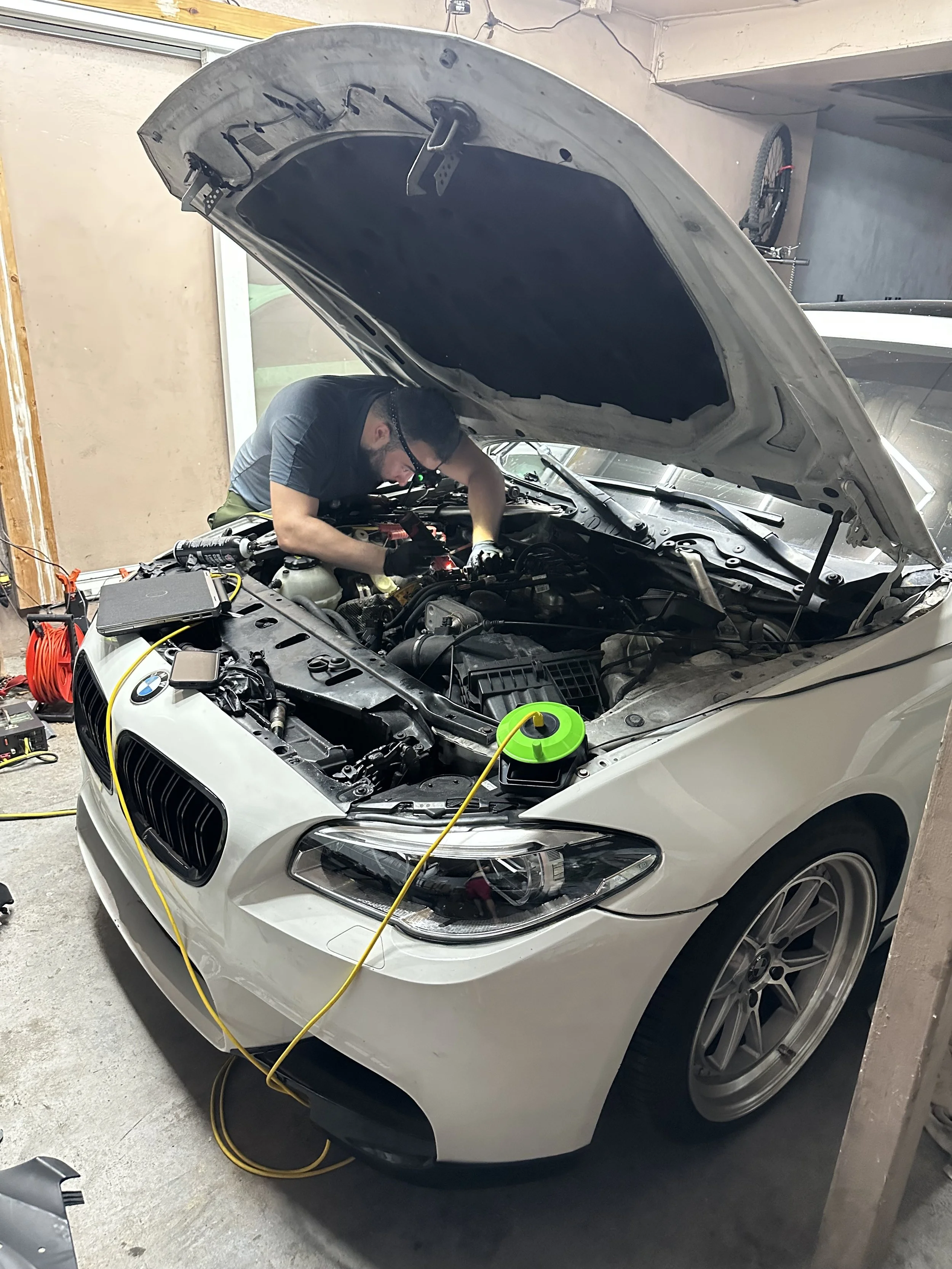 A man working under the open hood of a white BMW in a garage, surrounded by tools and equipment.