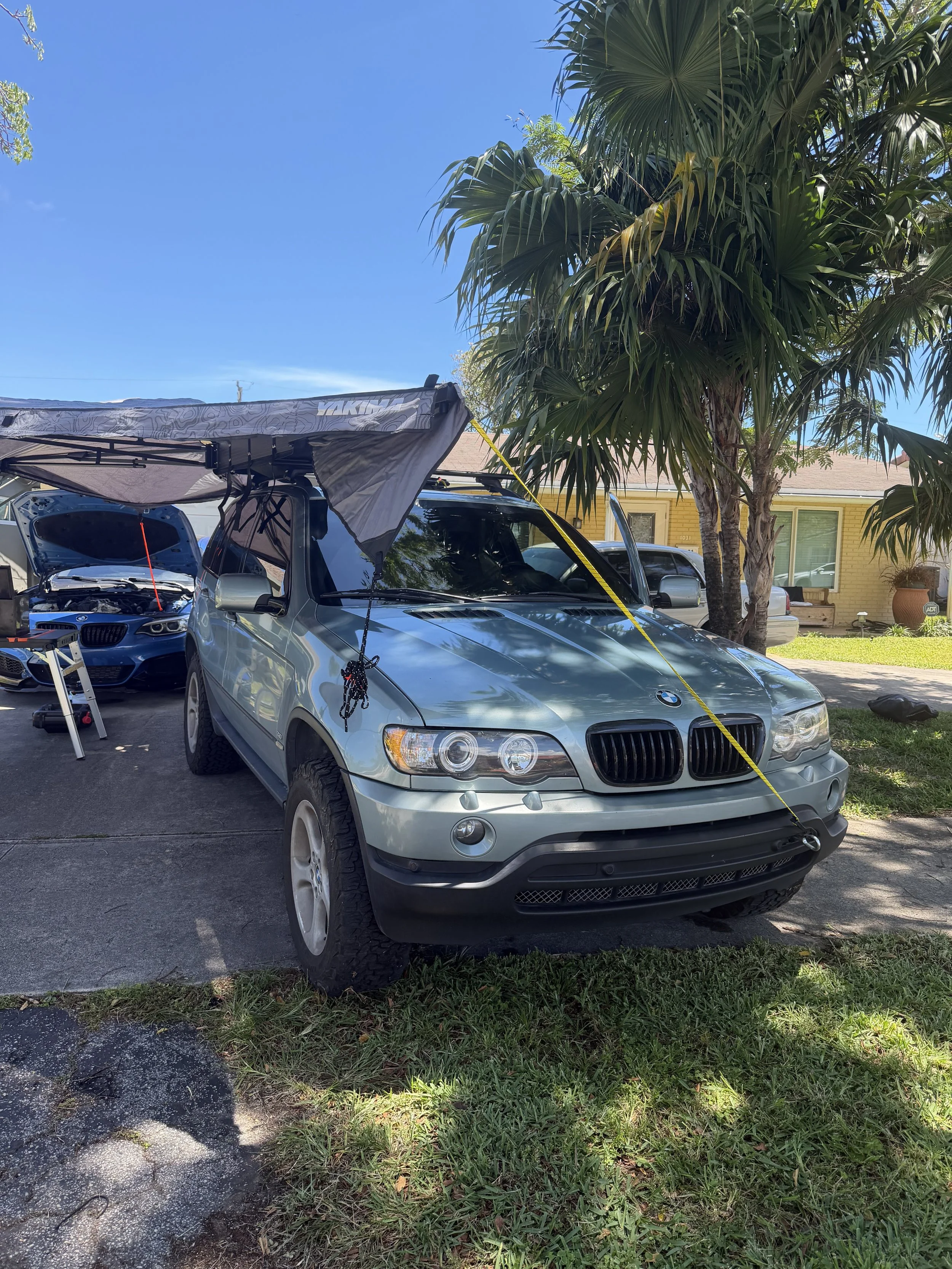 A silver BMW parked in a driveway with the hood open and a canopy extending from the car. There is a large palm tree and a yellow house in the background, and a blue sky overhead.