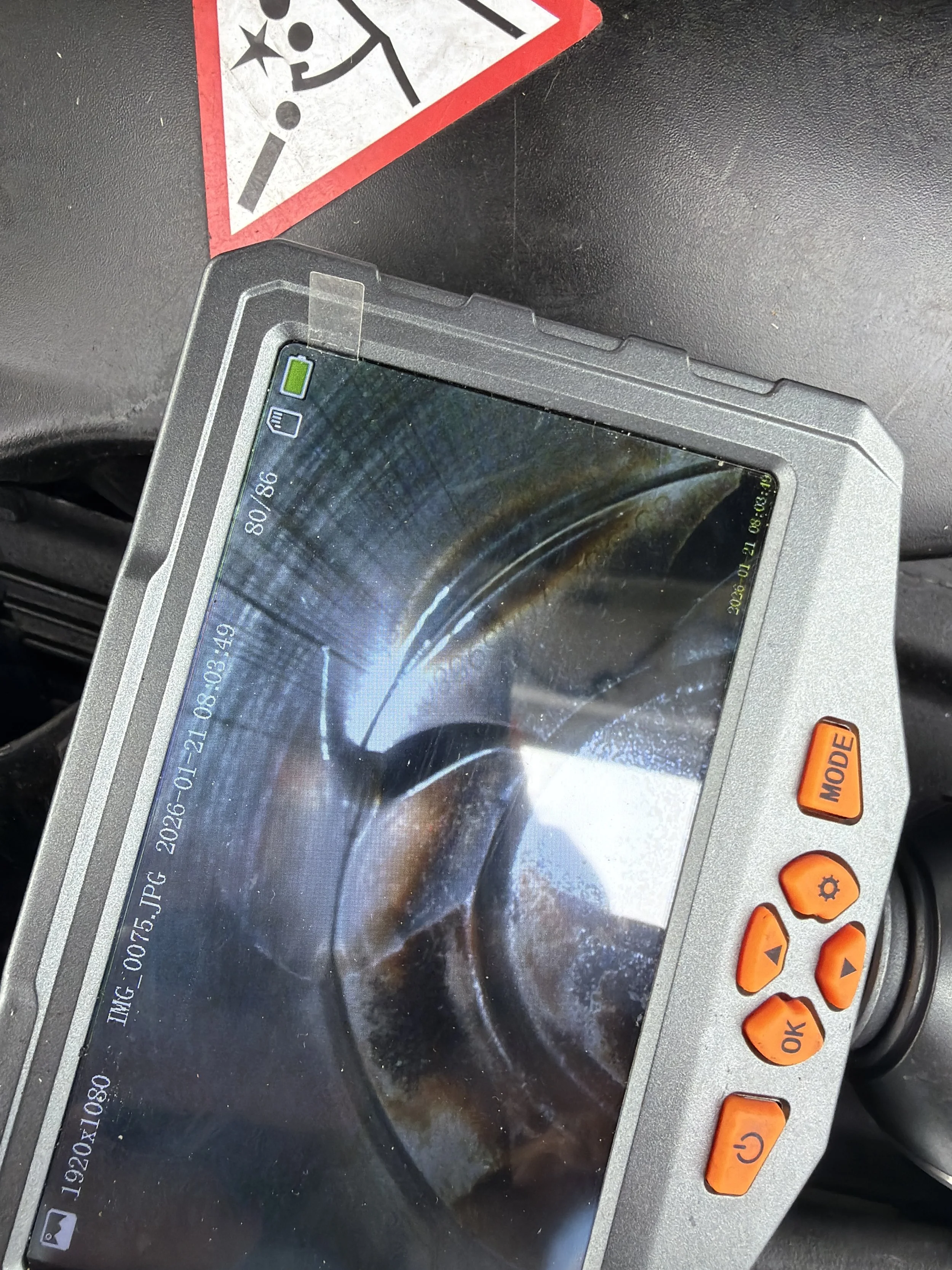 A digital device with a cracked screen and orange buttons, situated on a black surface with a warning sticker in the upper left corner.
