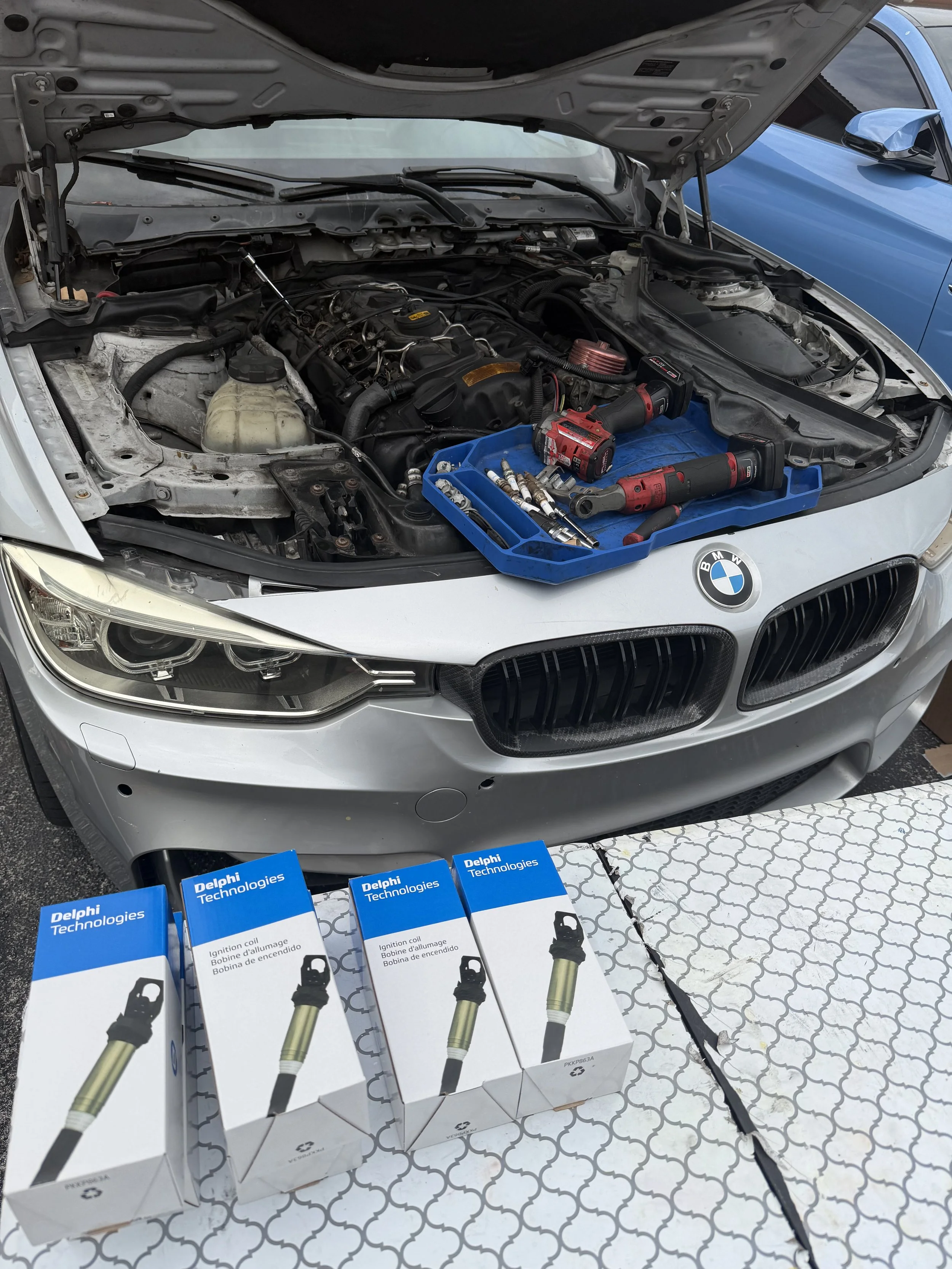 Open car hood with tools and spark plug ignition coils displayed on the front grille of a silver BMW vehicle.