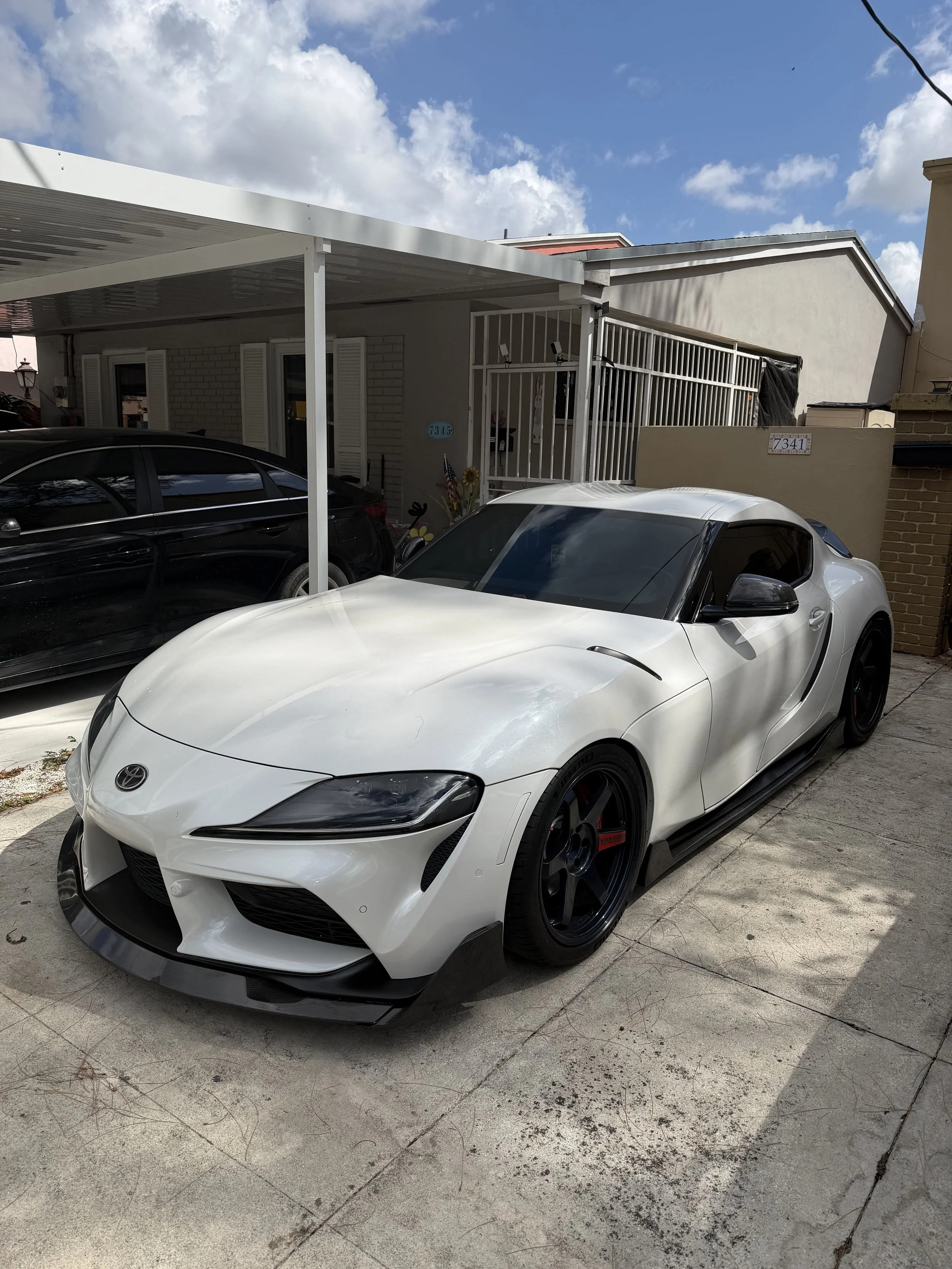 A white sports car with black wheels parked on a driveway in front of a house, with another black car partially visible beside it. The house has a covered porch and a gate, with a blue house number sign visible.