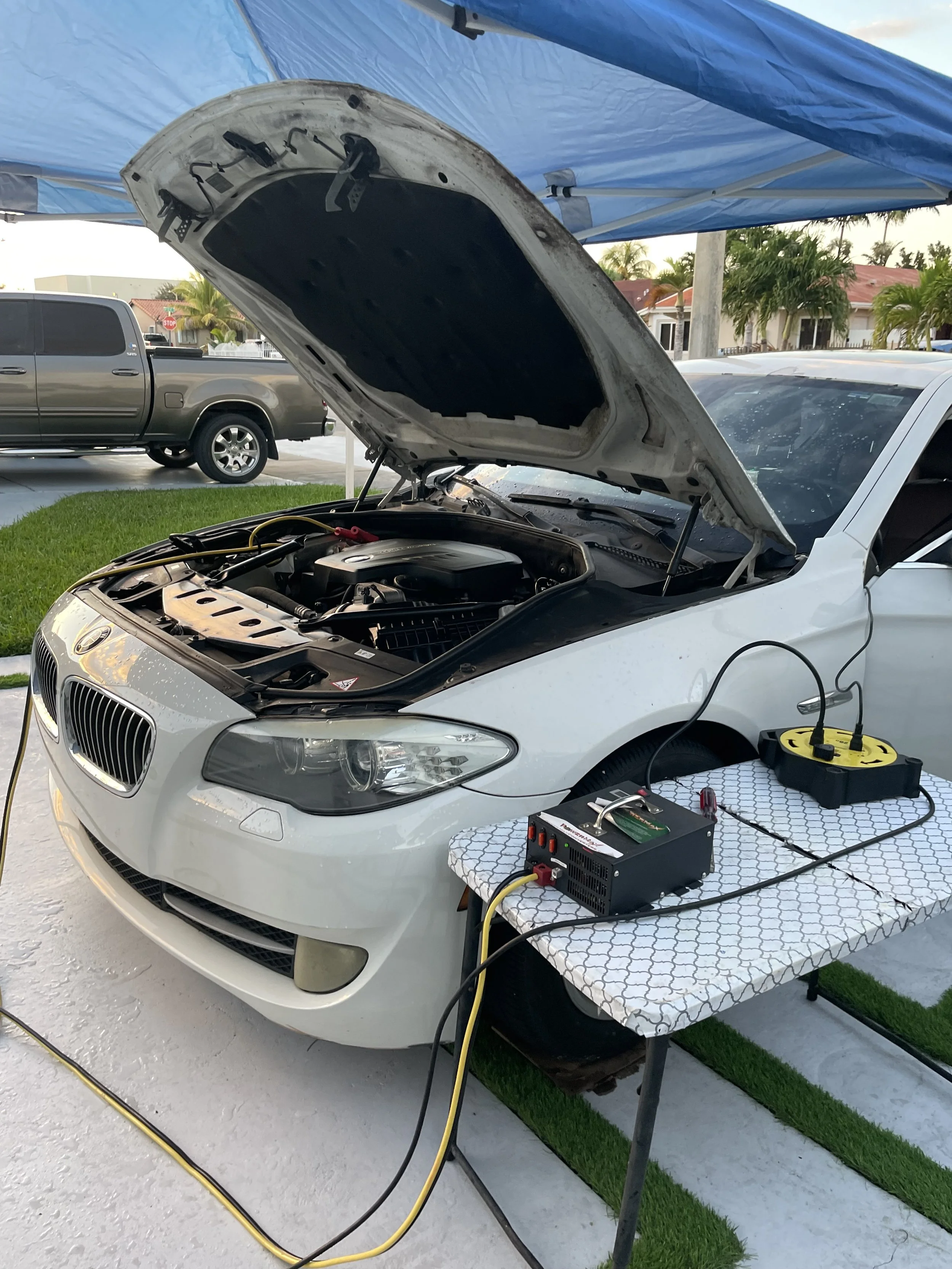 A white car with its hood open parked on a concrete driveway. Diagnostic equipment and wires are connected to the engine, with testing and diagnostic devices on a nearby table. Other vehicles and residential houses are visible in the background.