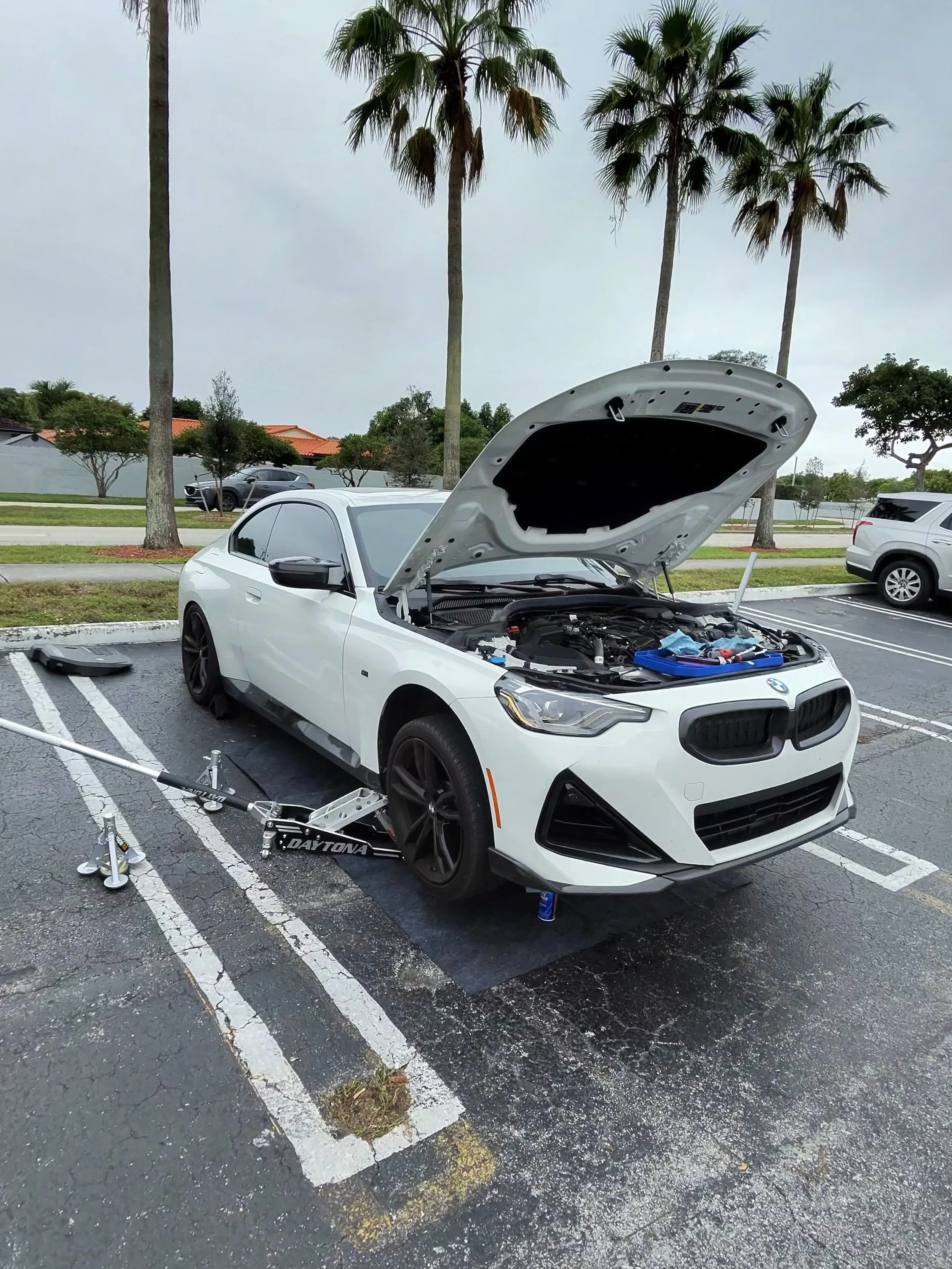 A white sports car with its hood open parked in an outdoor parking lot on a cloudy day. The front left tire is removed, with a jack and tools nearby. There are palm trees and a few other cars in the background.