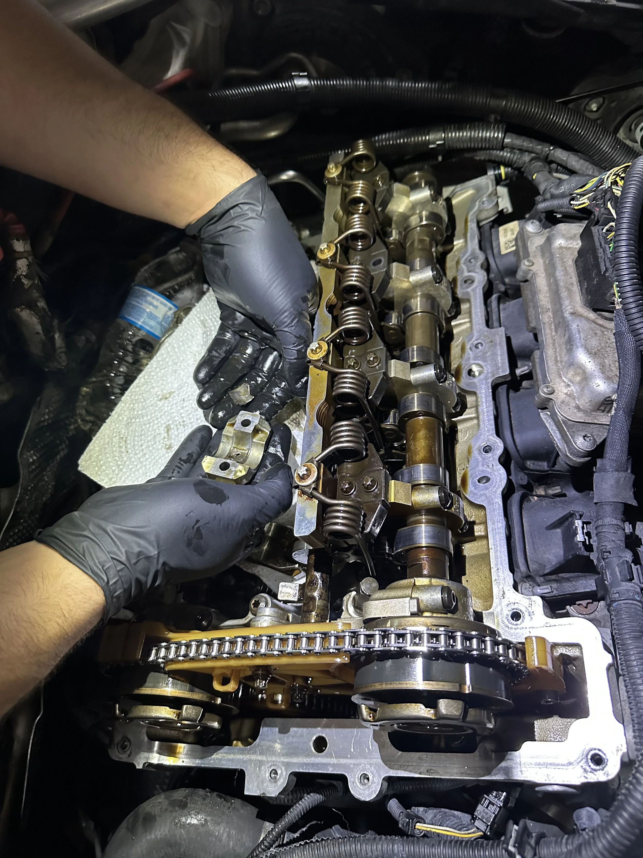A person wearing black gloves working on an engine, specifically on the valve train components such as rocker arms and valves, inside a car engine bay.