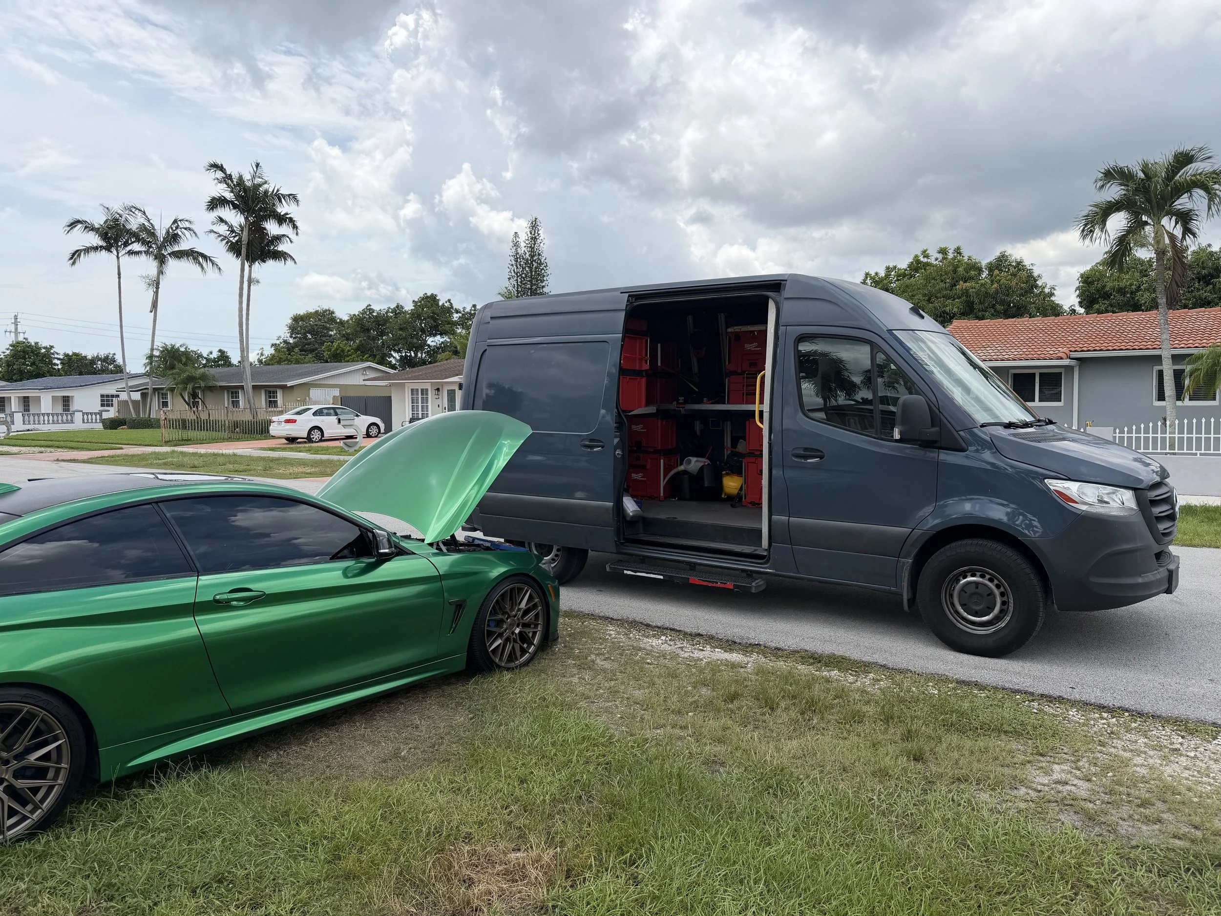 A black van with its side door open parked on a residential street, with a green sports car with an open hood in front. There are palm trees and houses in the background, with cloudy skies overhead.