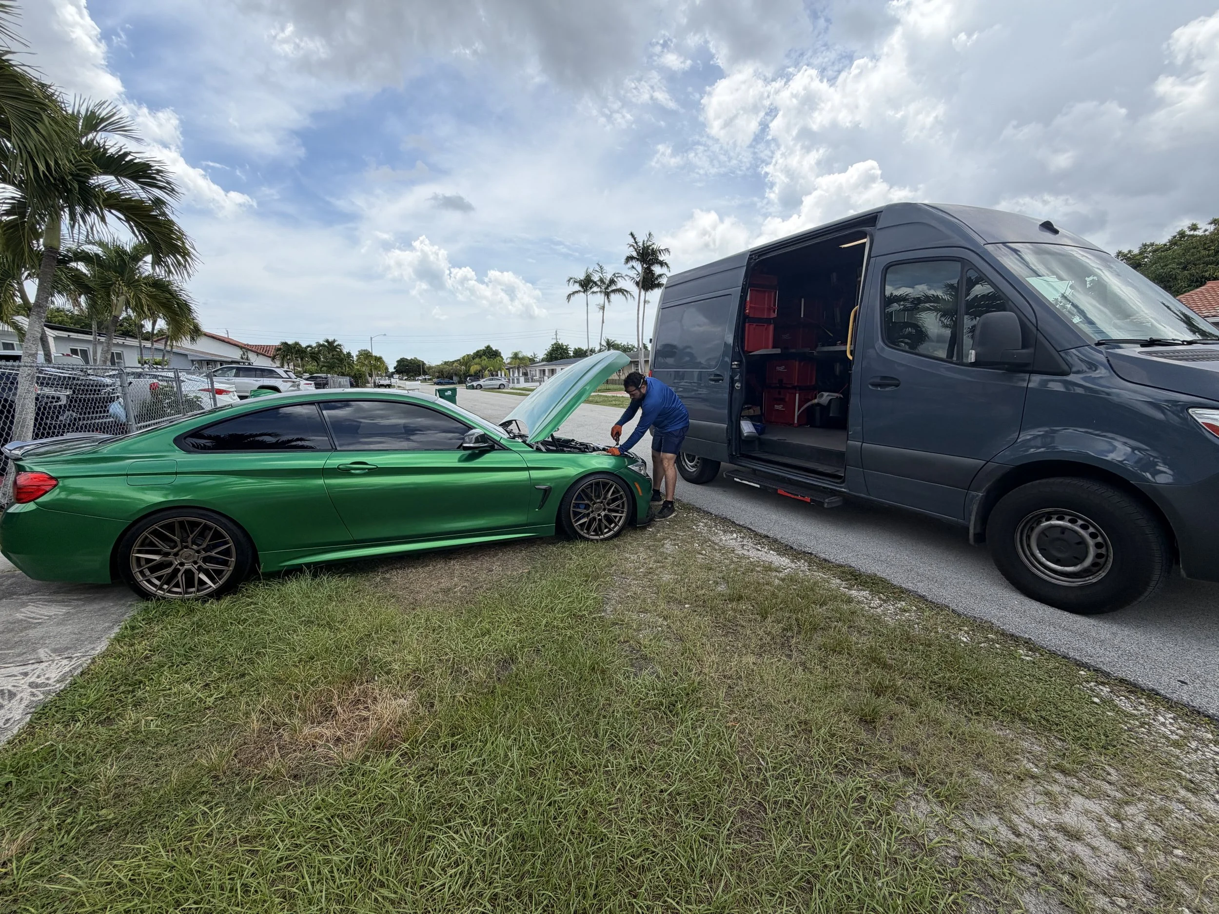 A man working on a green sports car with an open hood next to a large gray delivery van parked on the side of a residential street. There are palm trees and houses in the background under a partly cloudy sky.