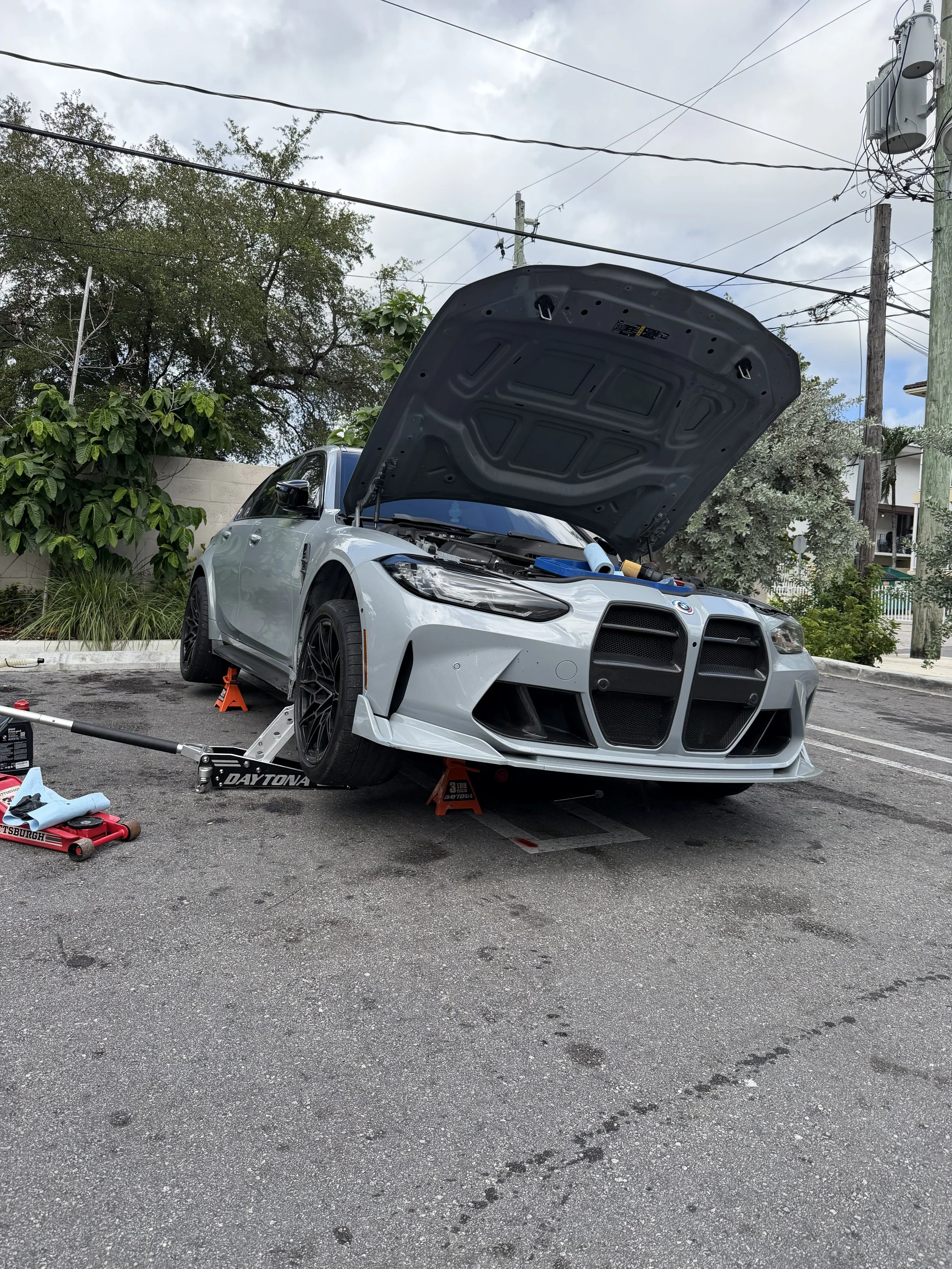 A silver sports car with an open hood, supported by jack stands, parked in an outdoor area with tools and equipment around, and trees and power lines in the background.