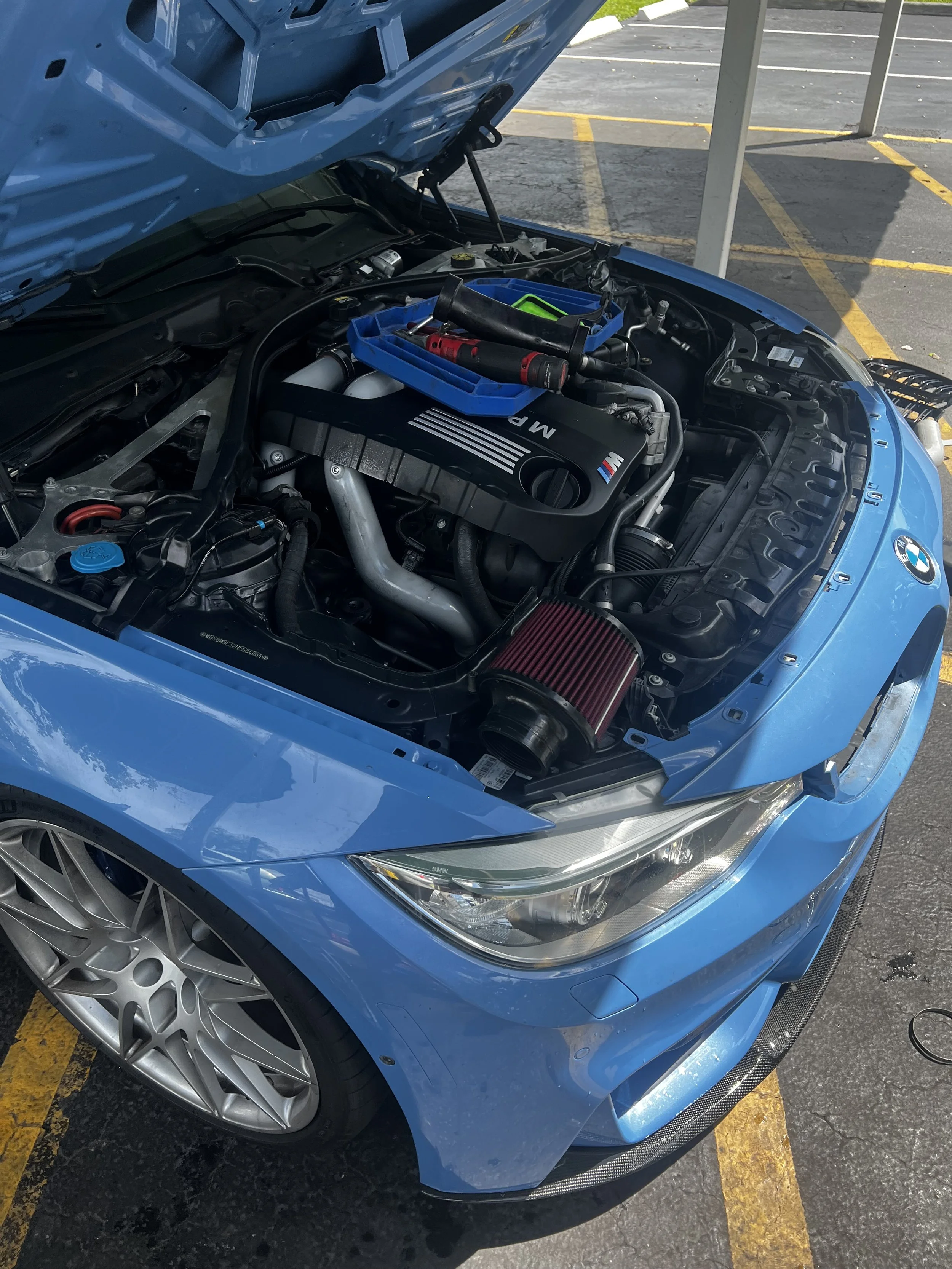 Blue sports car with its hood open in a parking lot, revealing the engine and tools placed on top of the engine.