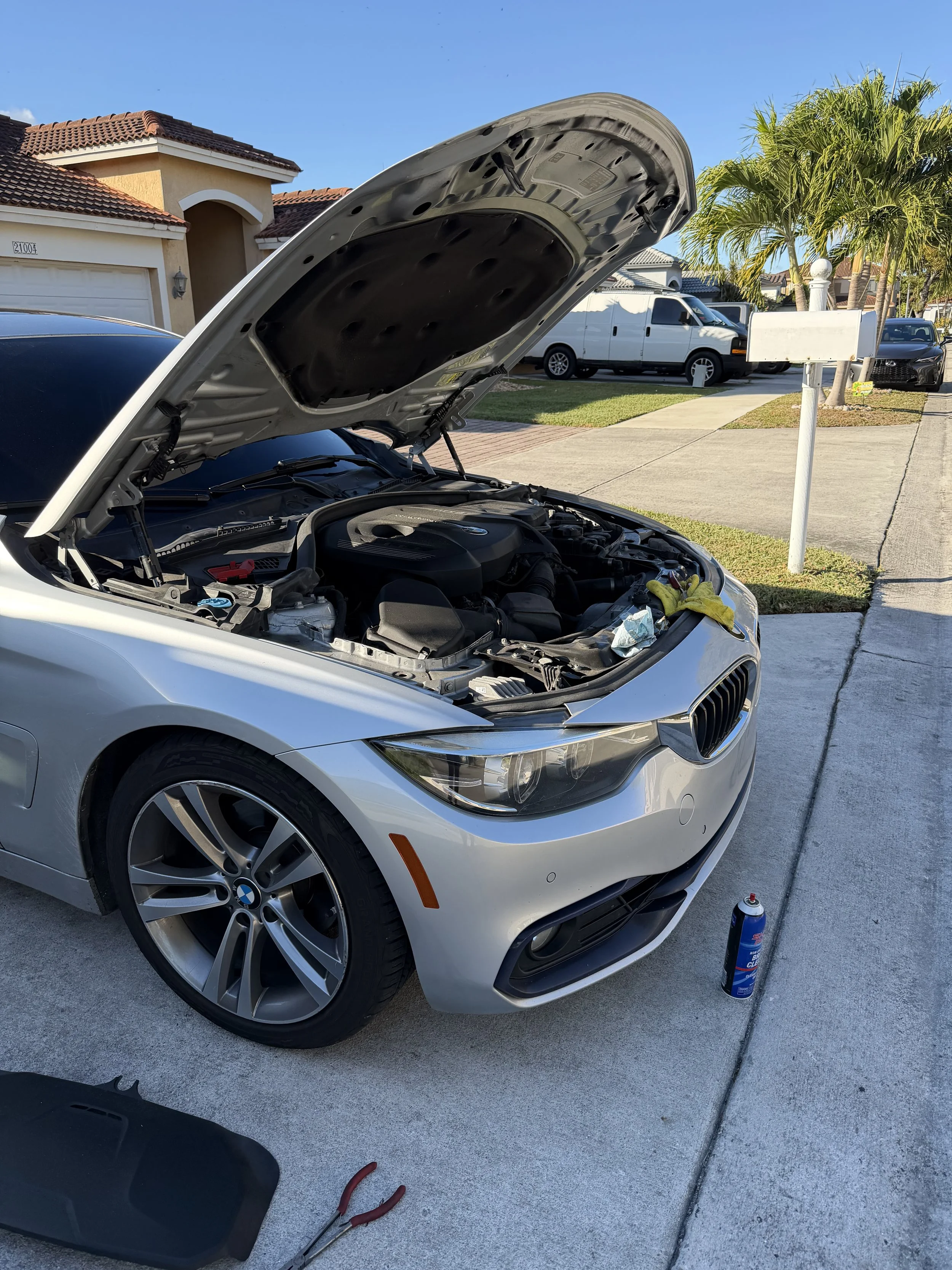 A silver BMW with its hood open, being serviced outside a residential house, with tools and a can of brake cleaner nearby.