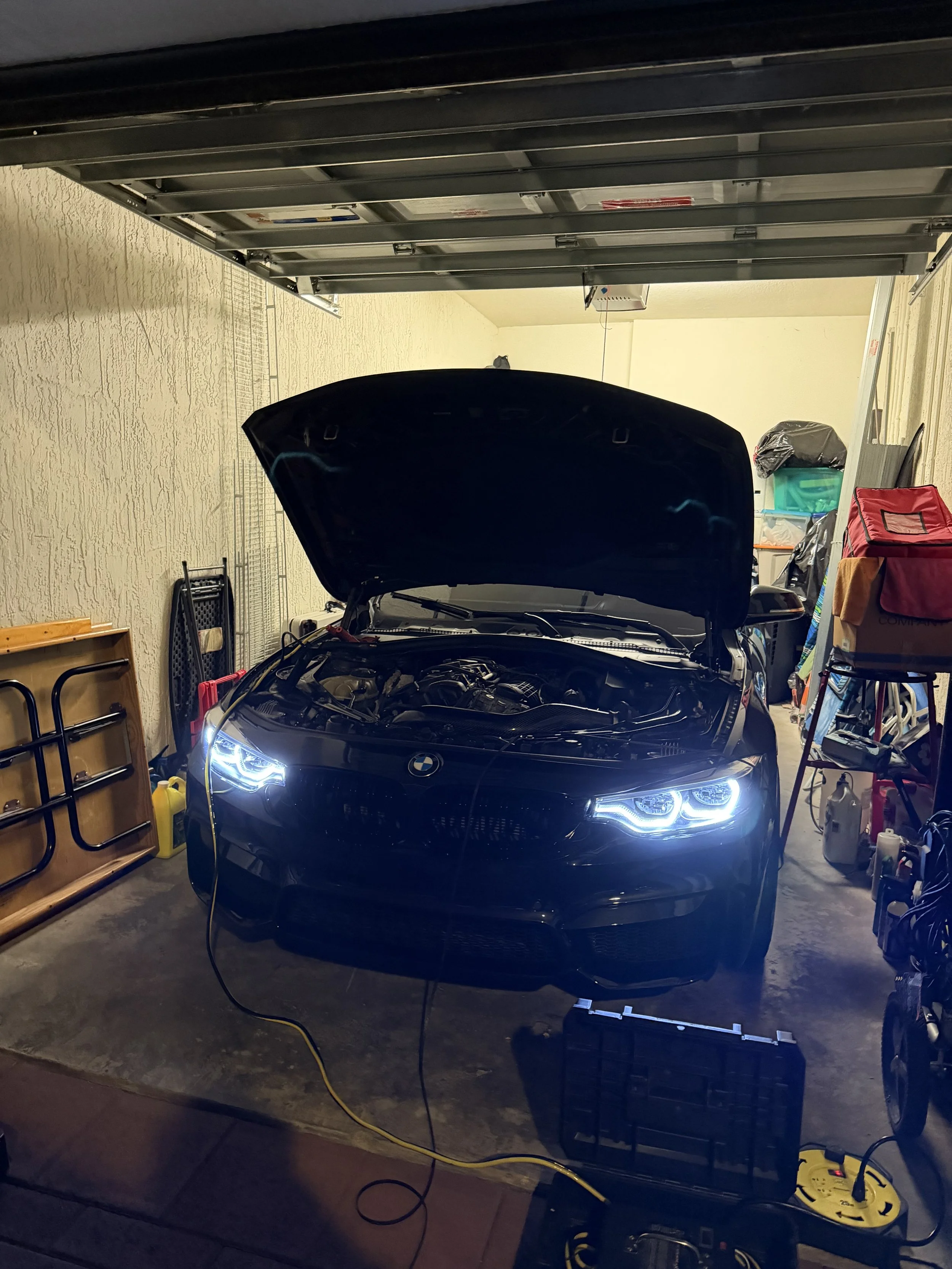 A black BMW with its hood open, parked inside a garage with cluttered shelves and a workbench. The headlights are on and tools and supplies are visible around the car.