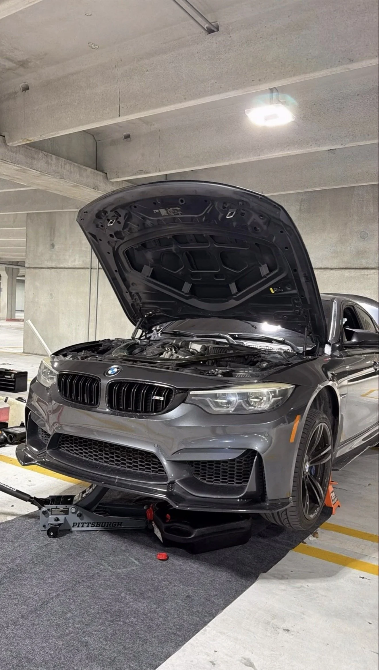 A black BMW car with its hood open, parked in an underground parking garage, with car maintenance tools and equipment nearby.