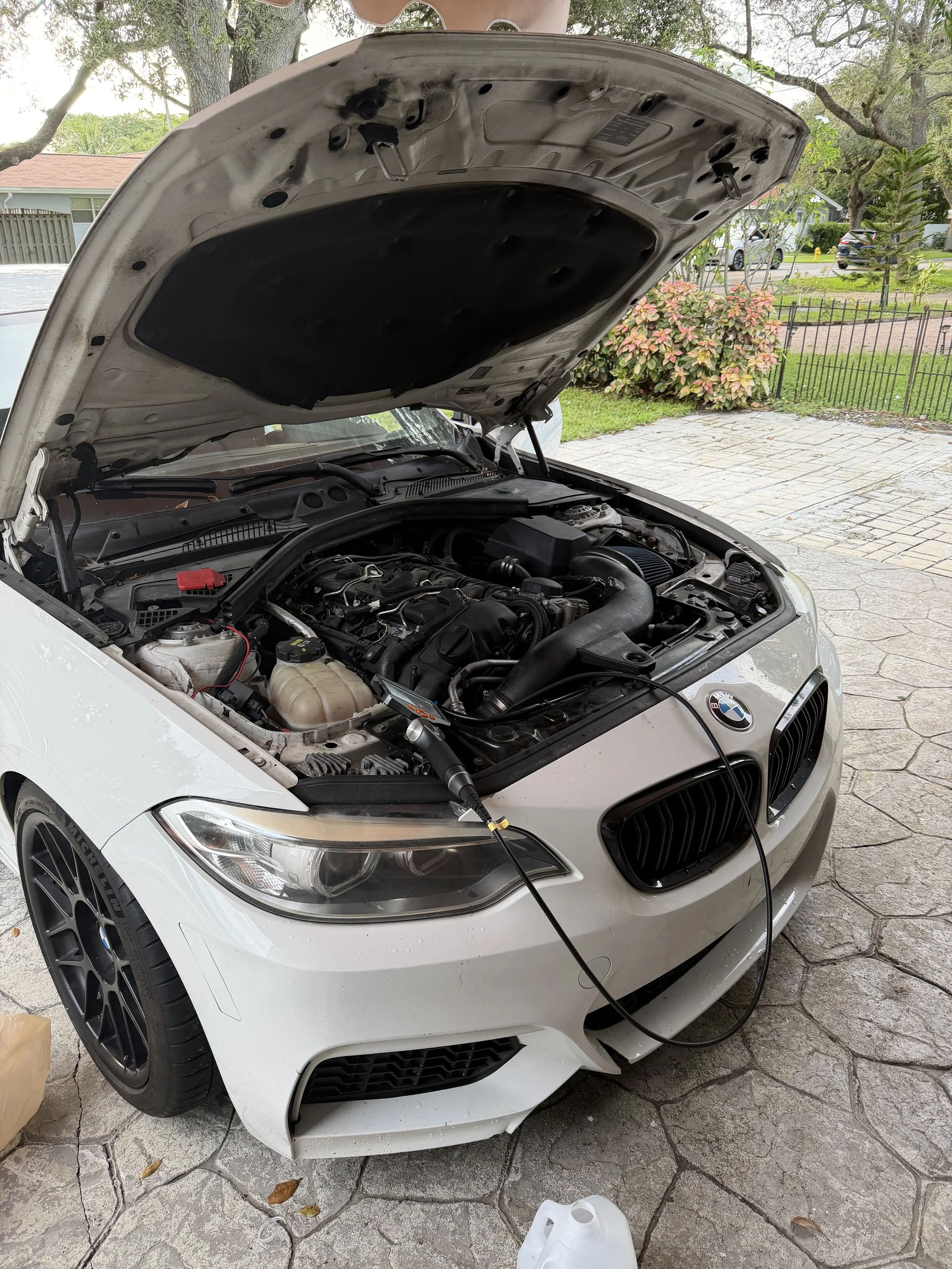 A white BMW car with its hood open, connected to a portable battery jump starter by cables, parked on a concrete driveway with a background of trees, shrubs, and houses.