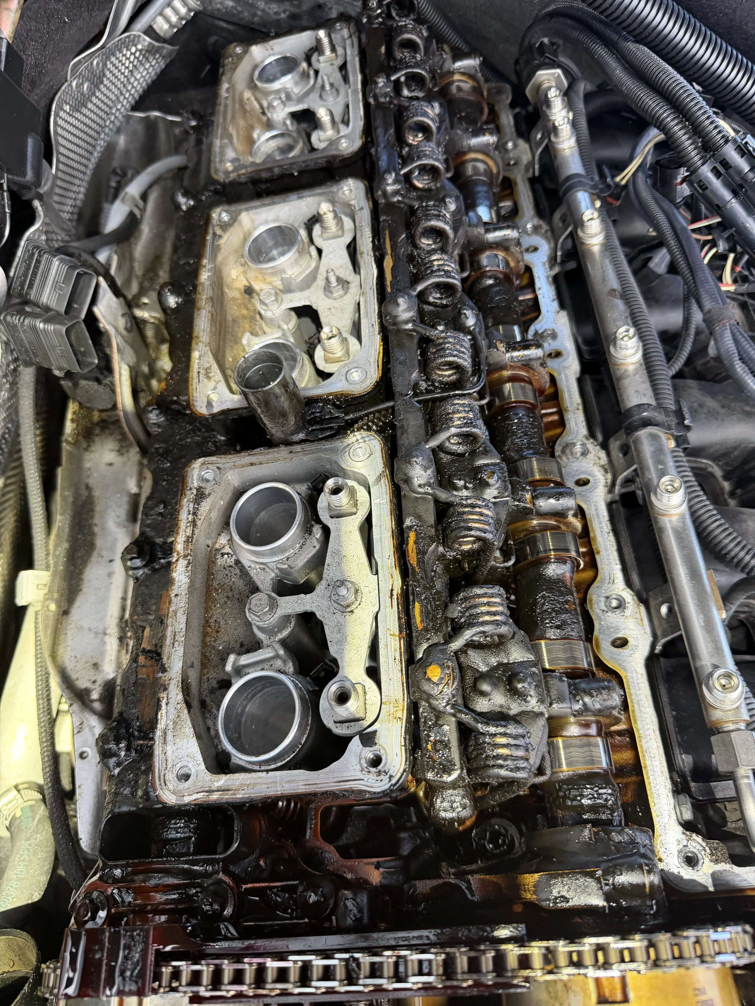 A disassembled car engine with the valve cover removed, exposing the camshafts, valve springs, and other internal components, all covered in oil and grime.