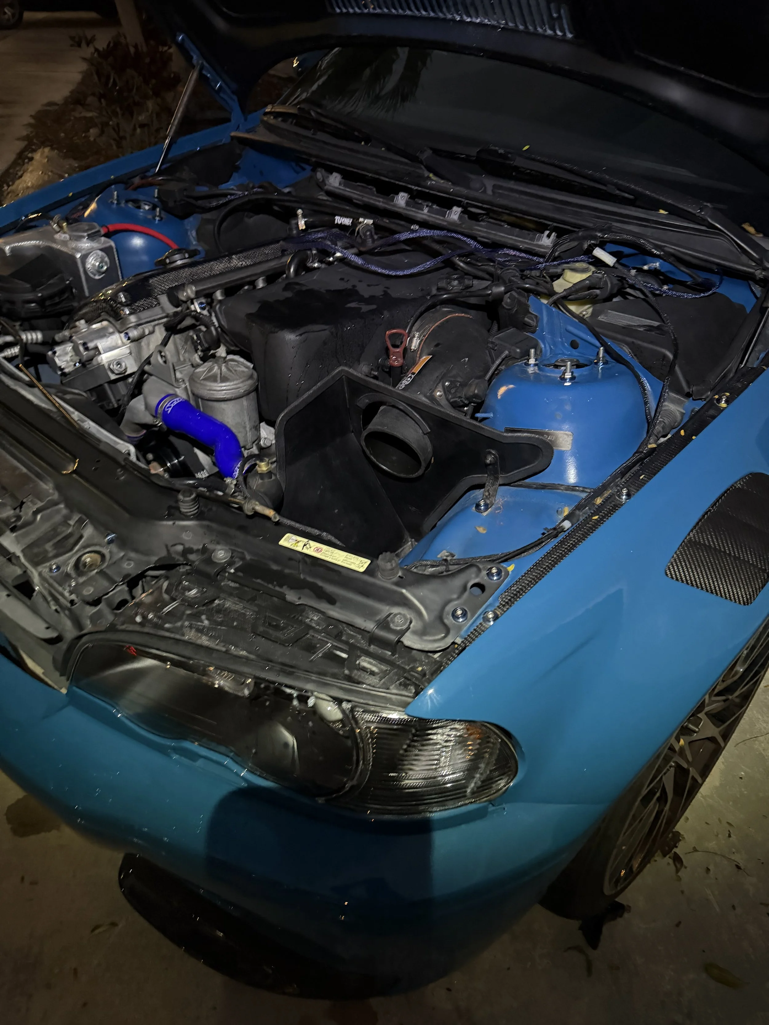 Close-up of a blue car with its hood open, revealing the engine bay, headlight, and front bumper.