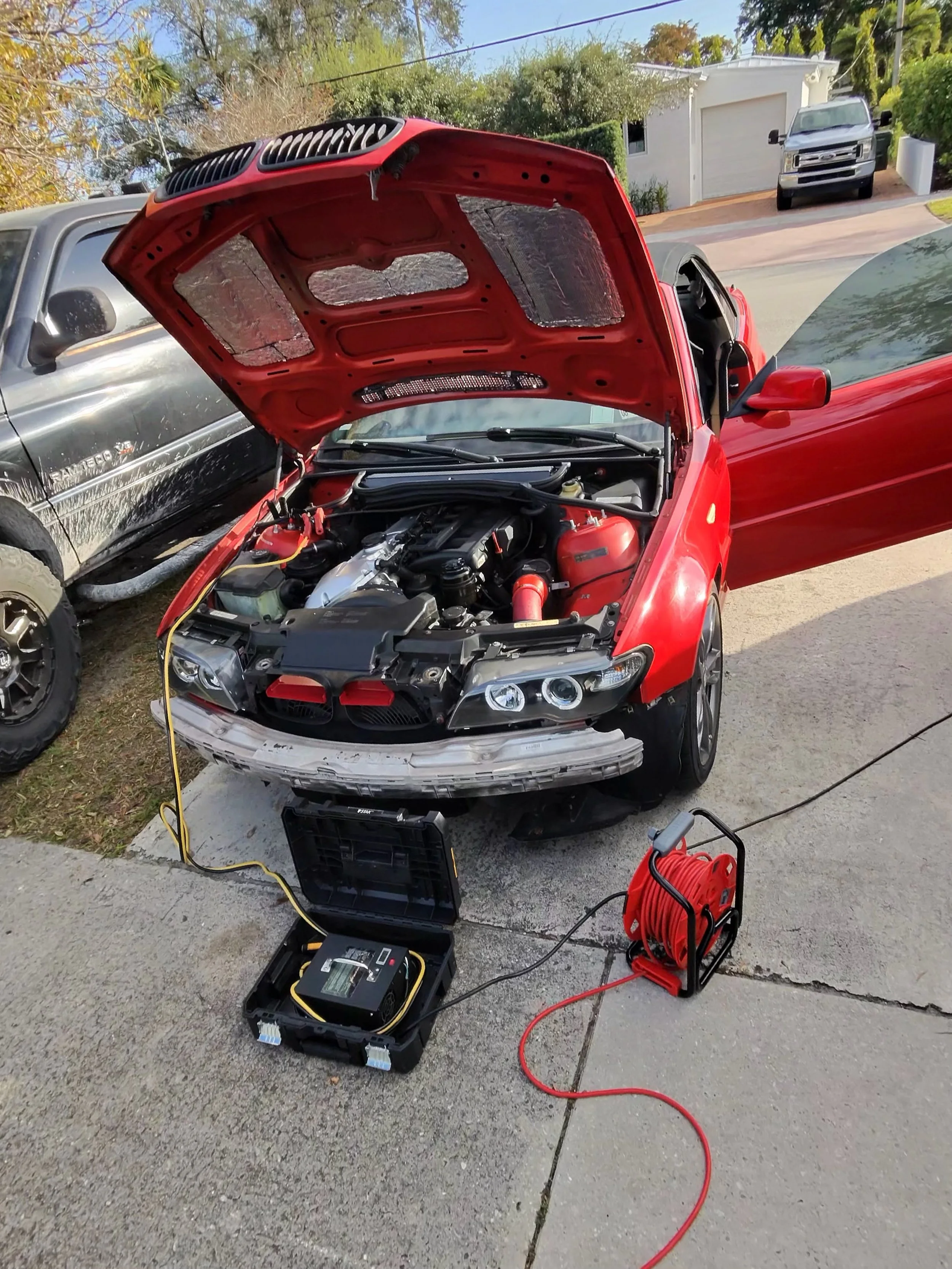 A red car with its hood open, being serviced with diagnostic equipment and a power tester on the driveway of a suburban home.
