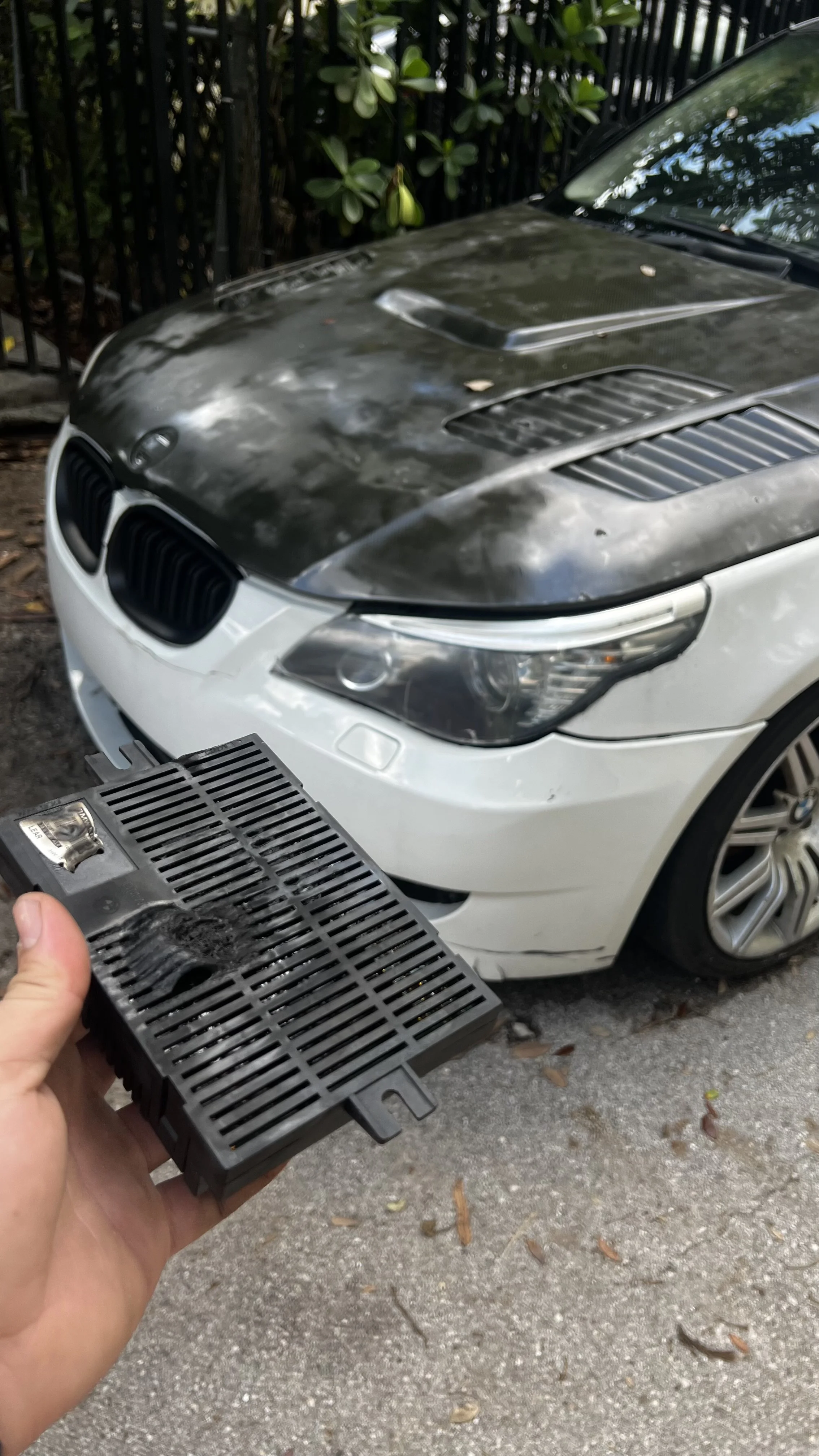 Person holding an engine control module in front of a damaged BMW car with a black hood, parked outdoors near a fence and trees.