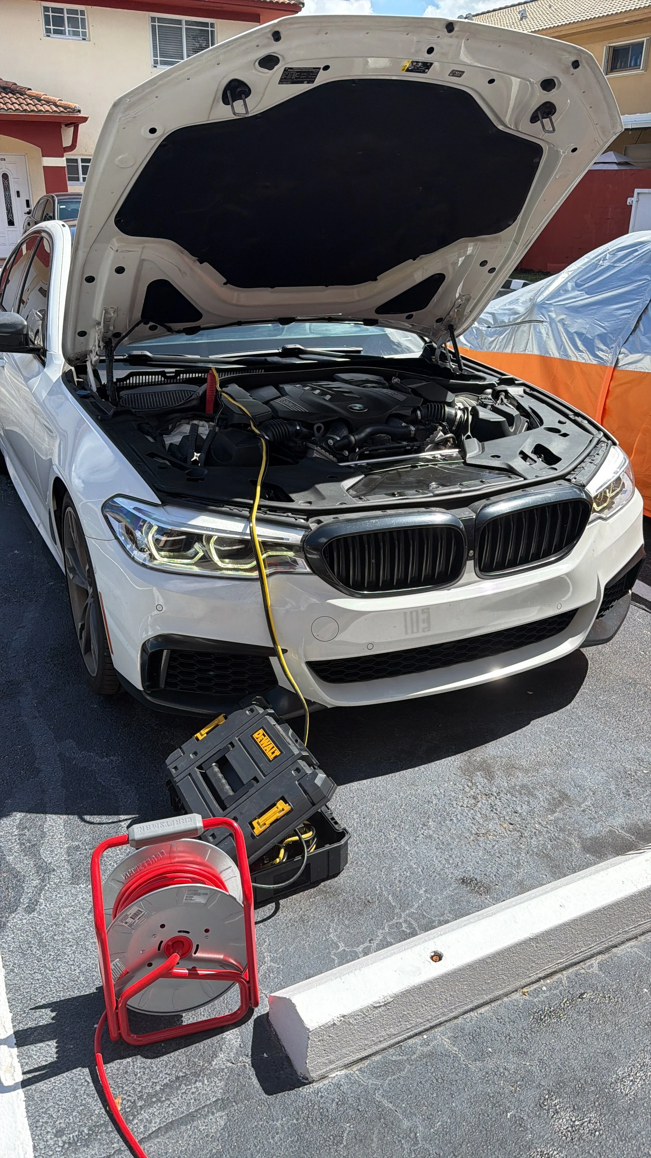 A white BMW car with its hood open, undergoing maintenance or repair in an outdoor parking area. Various tools and equipment are visible nearby, including a red extension cord reel, a black toolbox, and a cordless drill. Residential buildings are in 