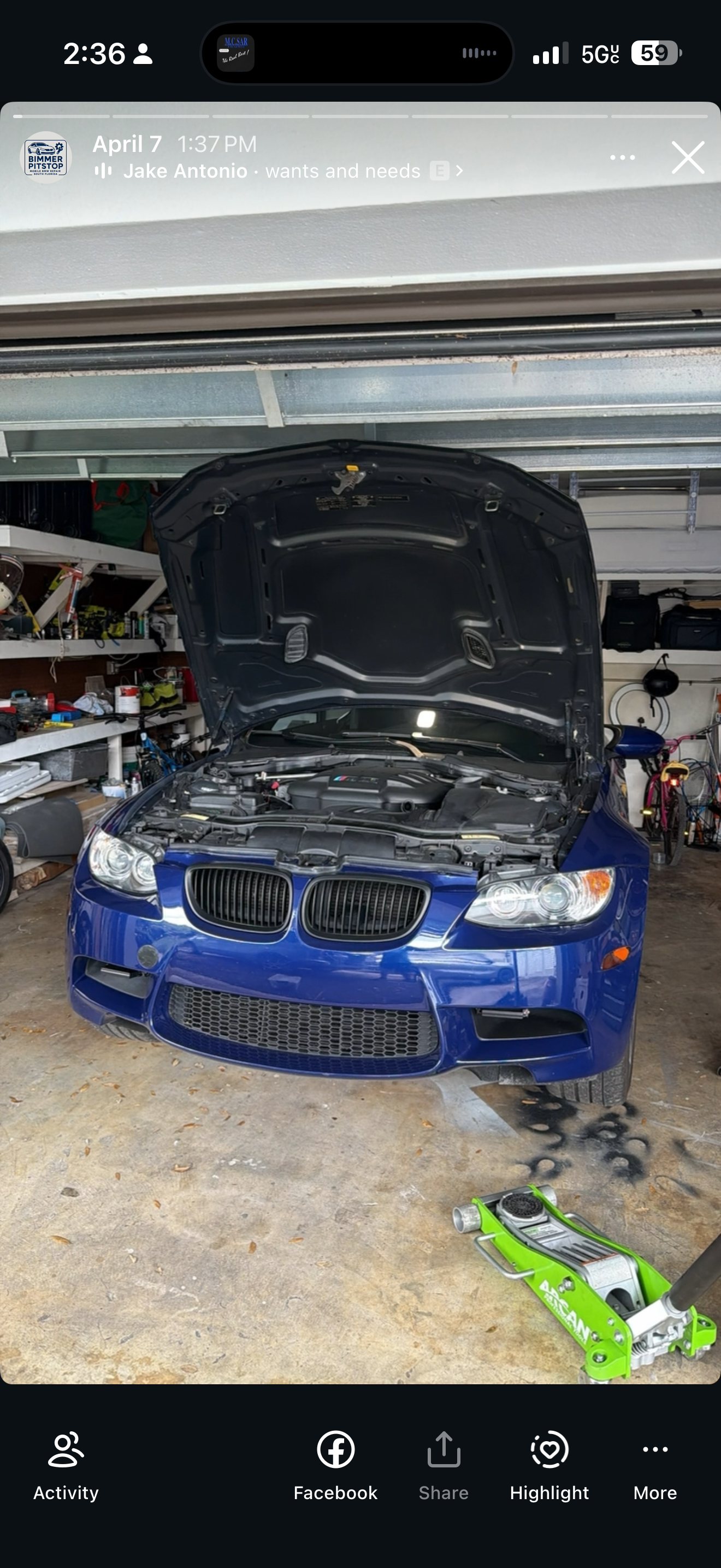 A blue BMW car with its hood open in a garage or workshop, surrounded by shelves with tools and equipment, and a green hydraulic floor jack in the foreground.