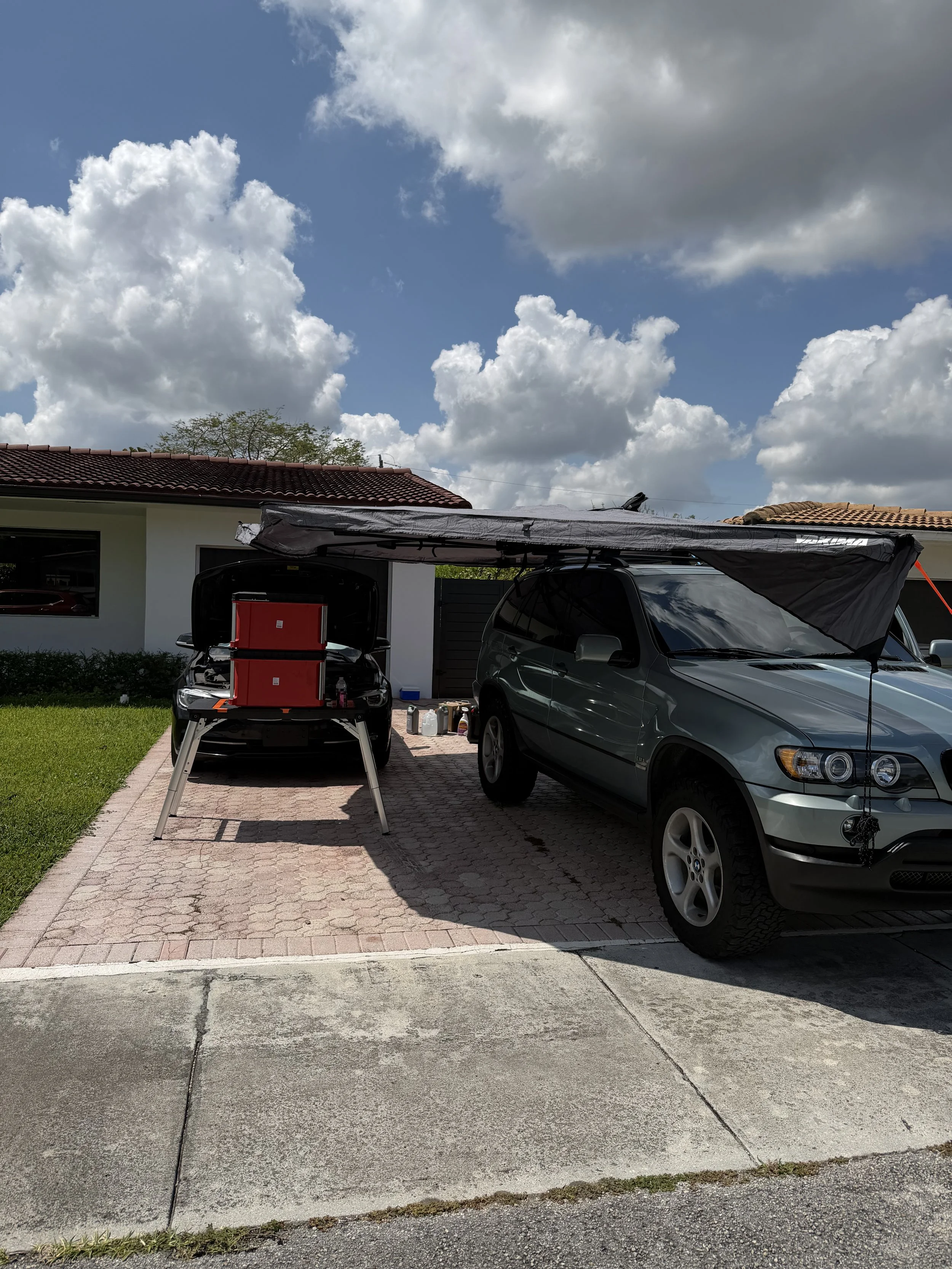 Two cars parked in a driveway in front of a house with a red-tiled roof, one with an open trunk showing equipment, and one with an extended canopy, under partly cloudy sky.