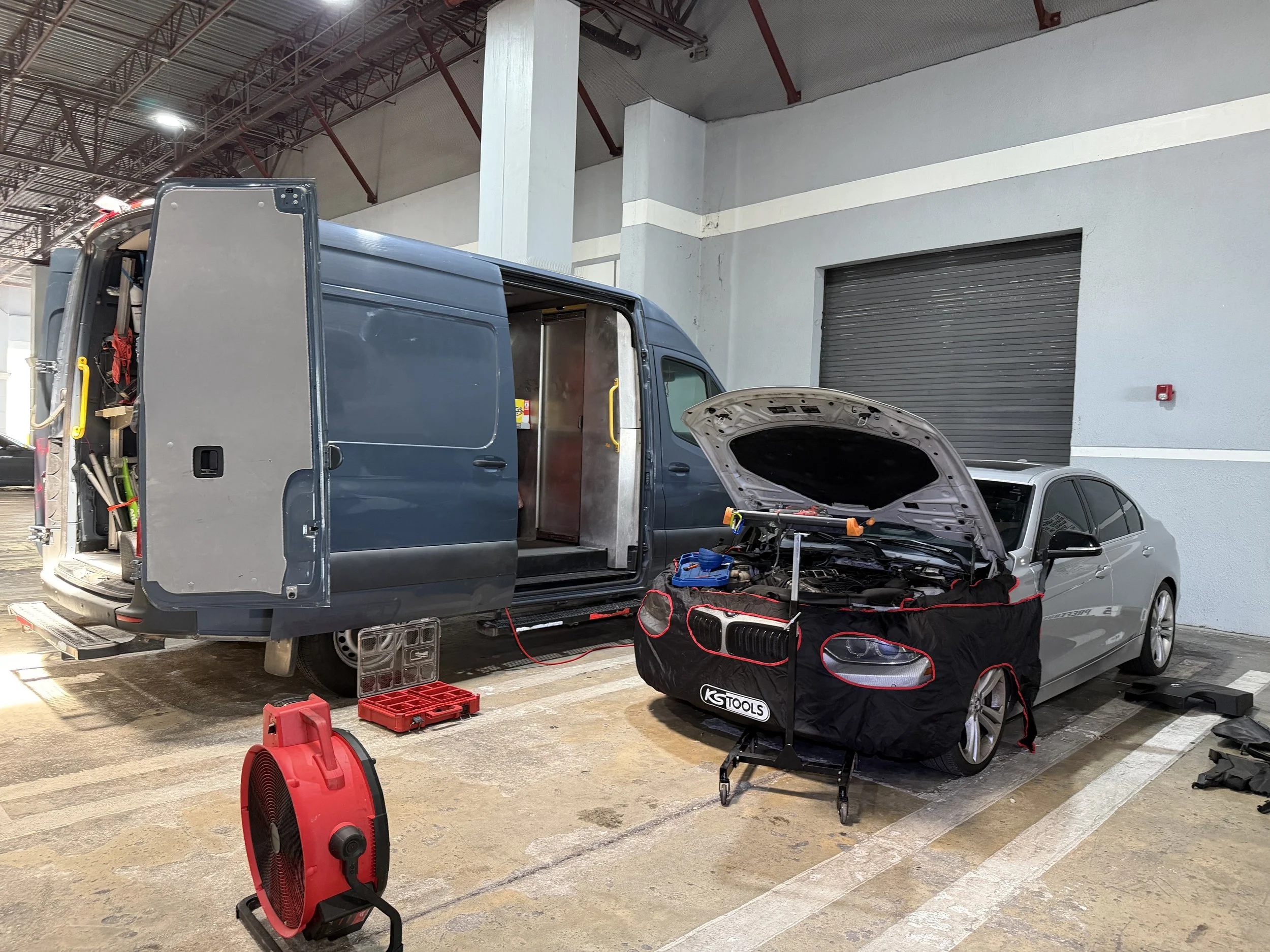 A service garage with a van and a sedan vehicle, both with open hoods, and tools and equipment on the floor and hanging inside the van. A red portable fan and a red heater are visible on the parking lot floor.