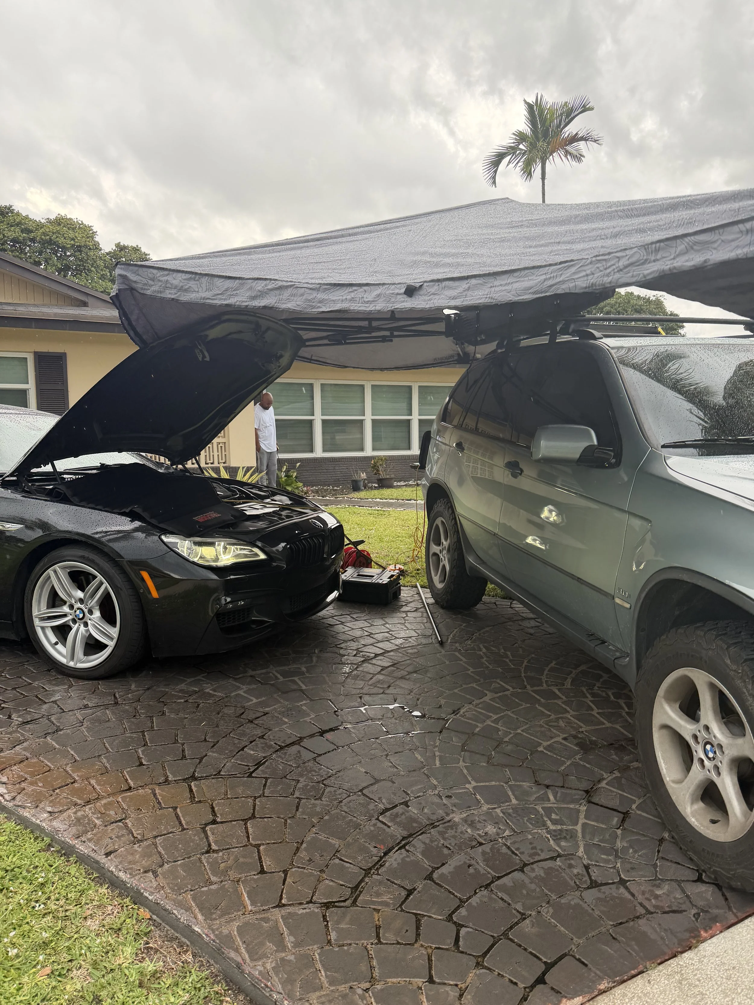 A black BMW with hood open parked on a cobblestone driveway, next to a gray vehicle with a canopy over both cars. A man stands near the house in the background, which has large windows and a tropical tree visible. The sky is cloudy.