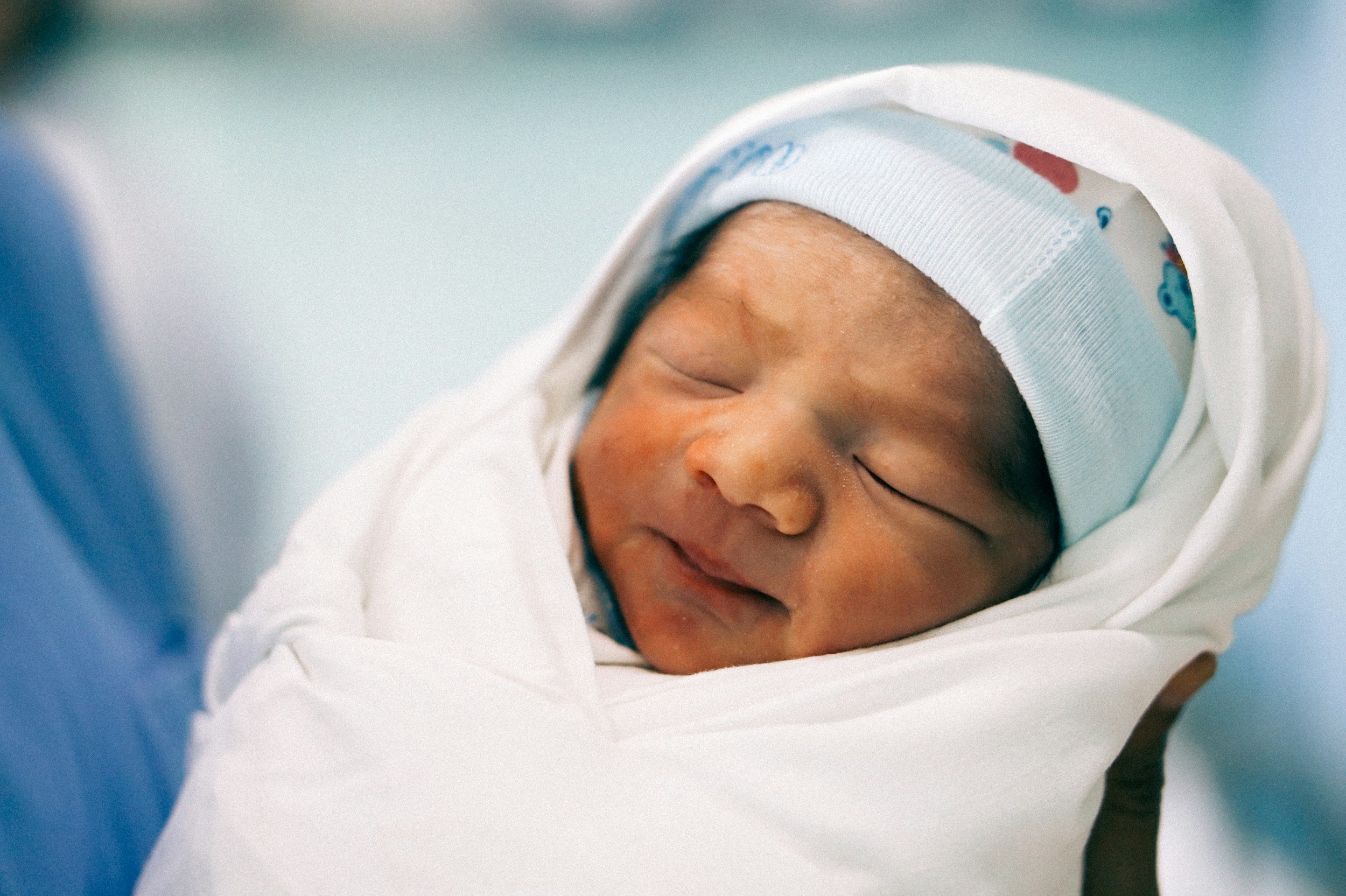 Close-up of a sleeping newborn baby with a beige skin tone, wrapped in a white blanket, wearing a white hat with blue and red designs. The baby is resting peacefully.