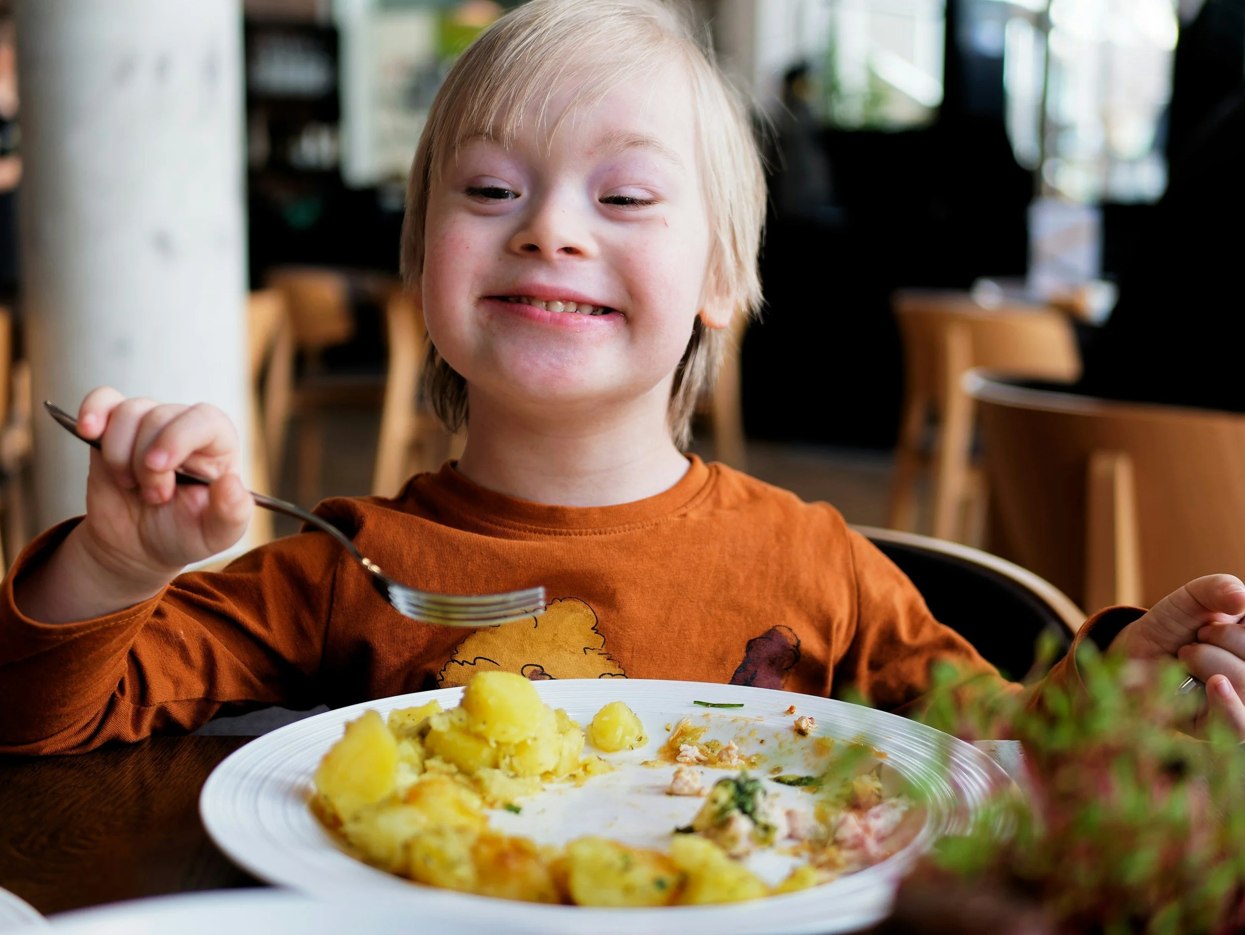 A smiling young boy with blonde hair in an orange shirt, sitting at a table with a plate of food, reaching with a spoon. The plate contains chunks of potatoes and other food, in a restaurant setting with chairs and large windows in the background.