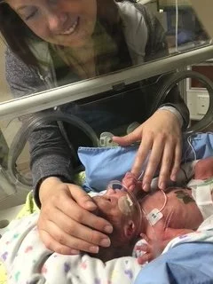 A woman gently holds the hand of a newborn baby in a hospital incubator, with the baby's face partially visible and medical wires attached.