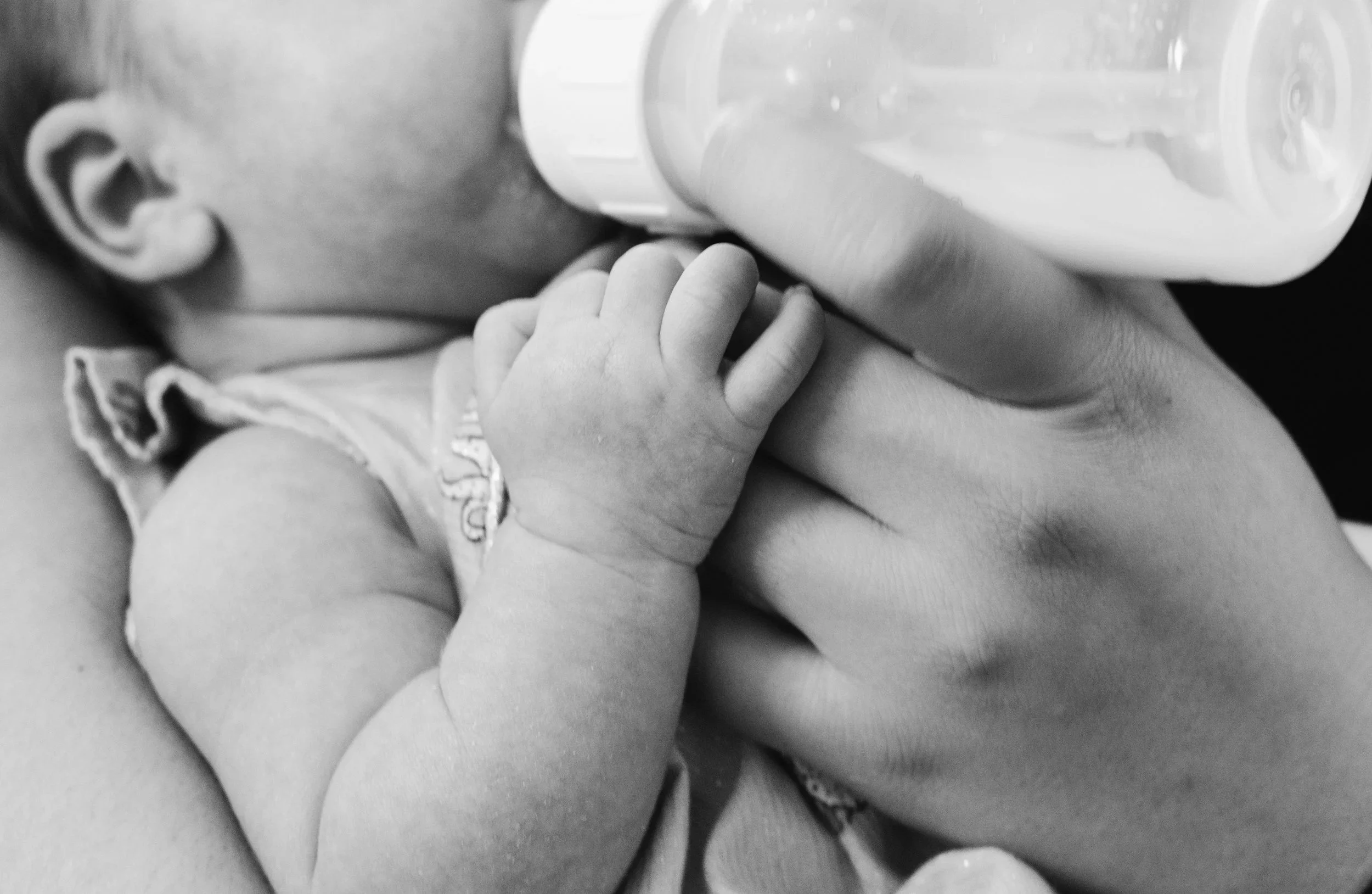 A close-up black-and-white photo of a baby's face with an eye and ear visible, holding a small bottle to their mouth with a hand.