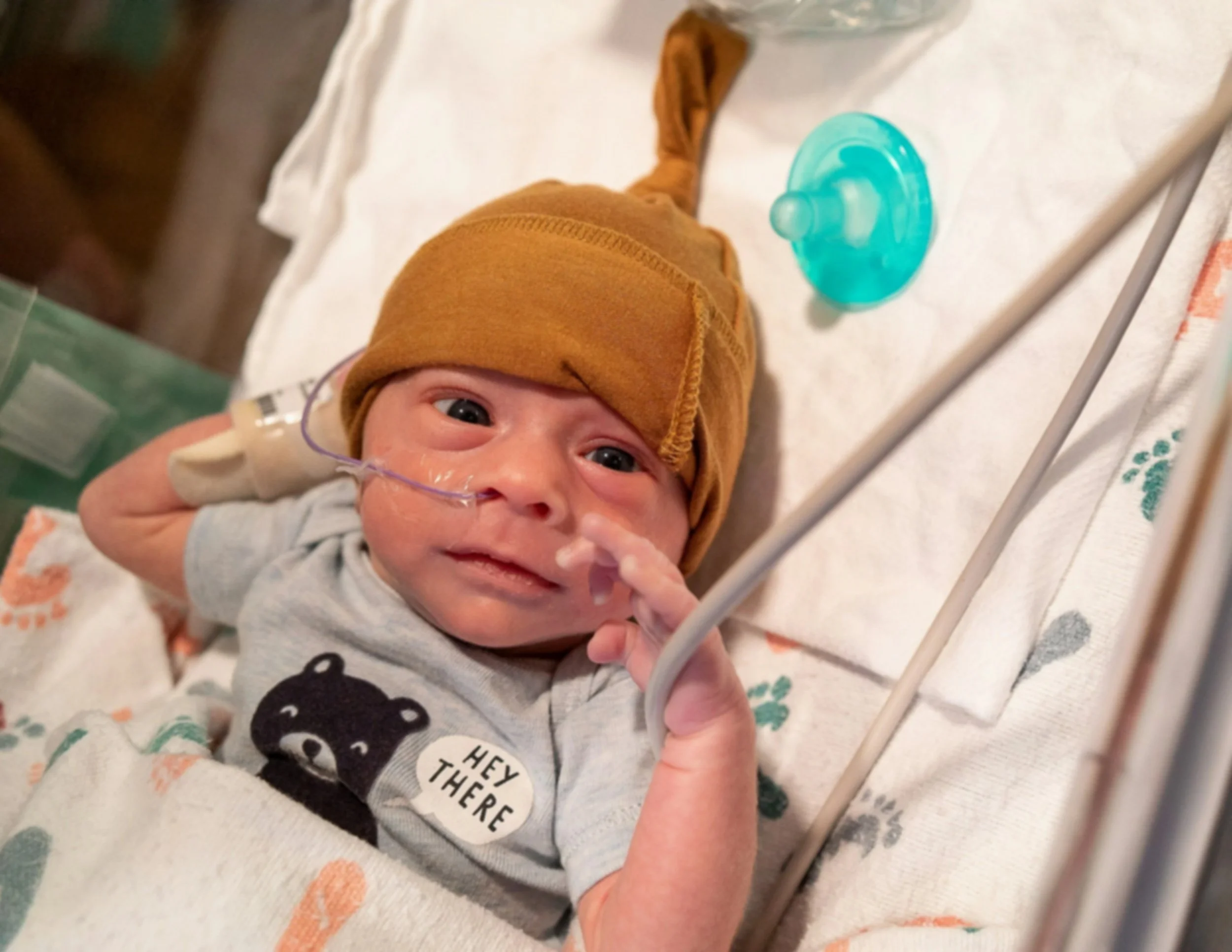 Close-up of a baby lying in an incubator with a nasal oxygen tube, wearing a brown hat and a onesie with bears and text, and looking at the camera.