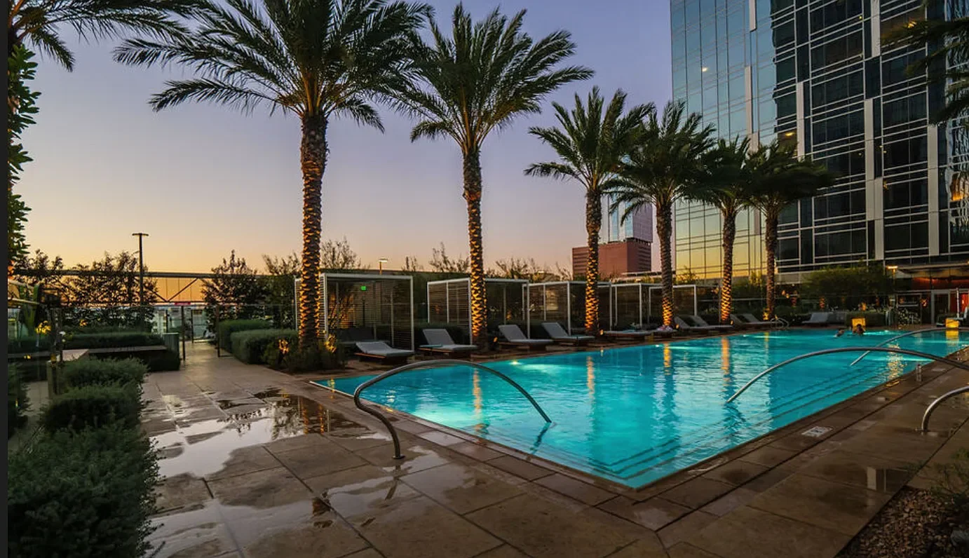 This is an outdoor hotel pool area at sunset with palm trees, lounge chairs, and a modern glass building in the background.