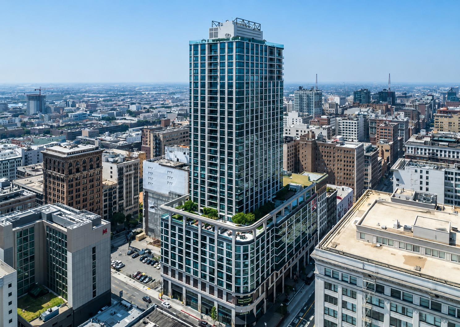 Aerial view of a city with a tall modern glass skyscraper featuring a rooftop garden, surrounded by other high-rise buildings and urban landscape under clear blue sky.