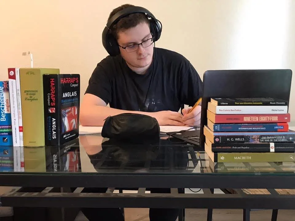 Jeune homme avec des lunettes, portant un casque audio, qui travaille à une table avec une collection de livres en anglais sur divers sujets (histoire, langue, littérature)