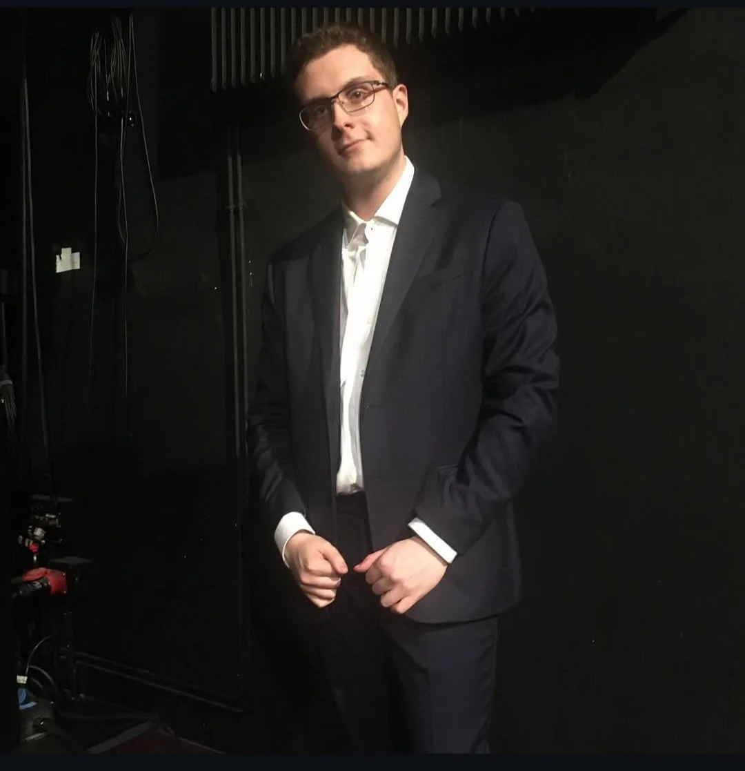 Jeune homme en costume noir, chemise blanche, portant des lunettes, posé de profil dans un endroit sombre.