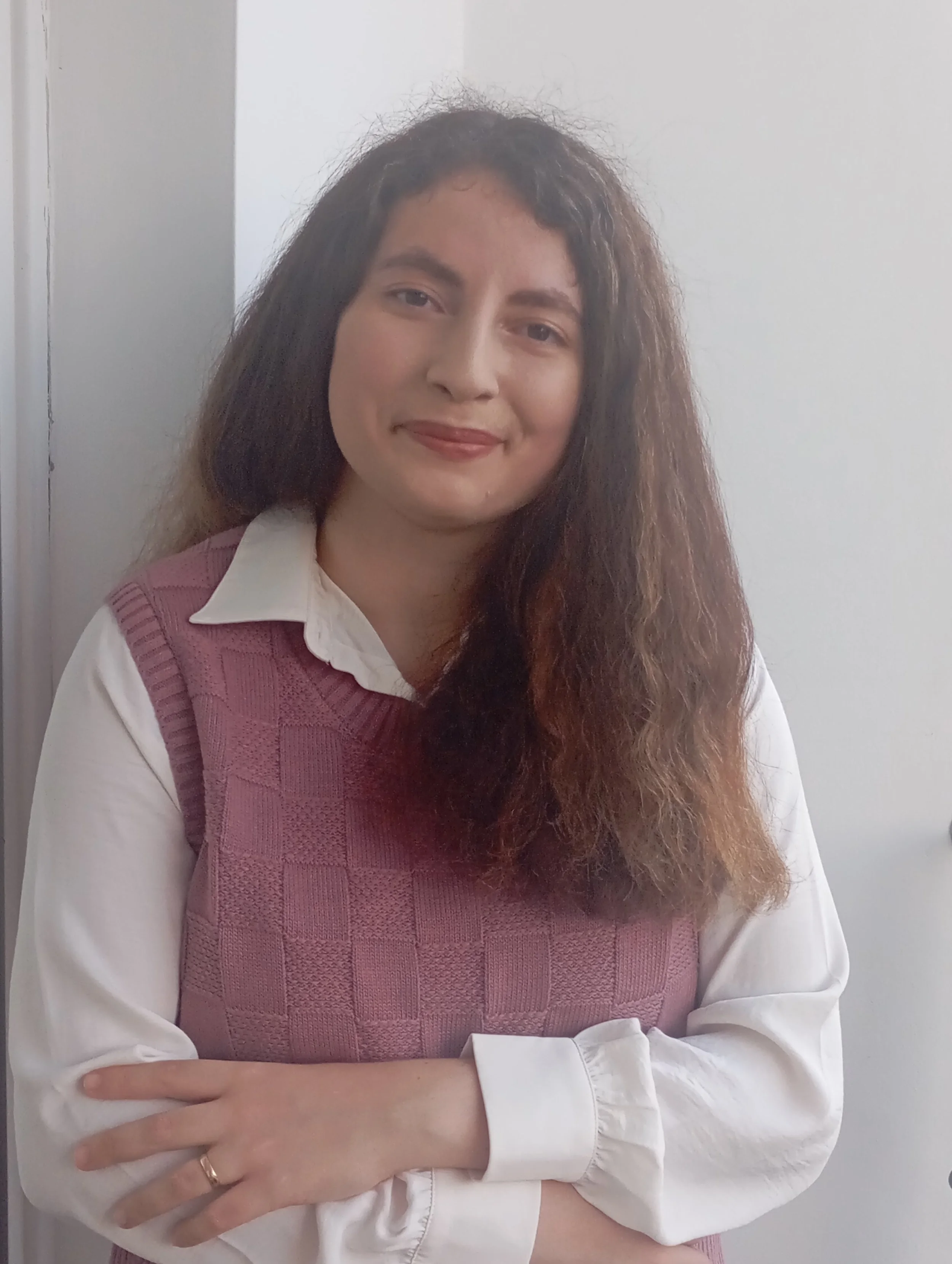 A woman with long, curly brown hair wearing a white shirt and pink vest, standing with arms crossed in front of a white and gray background.