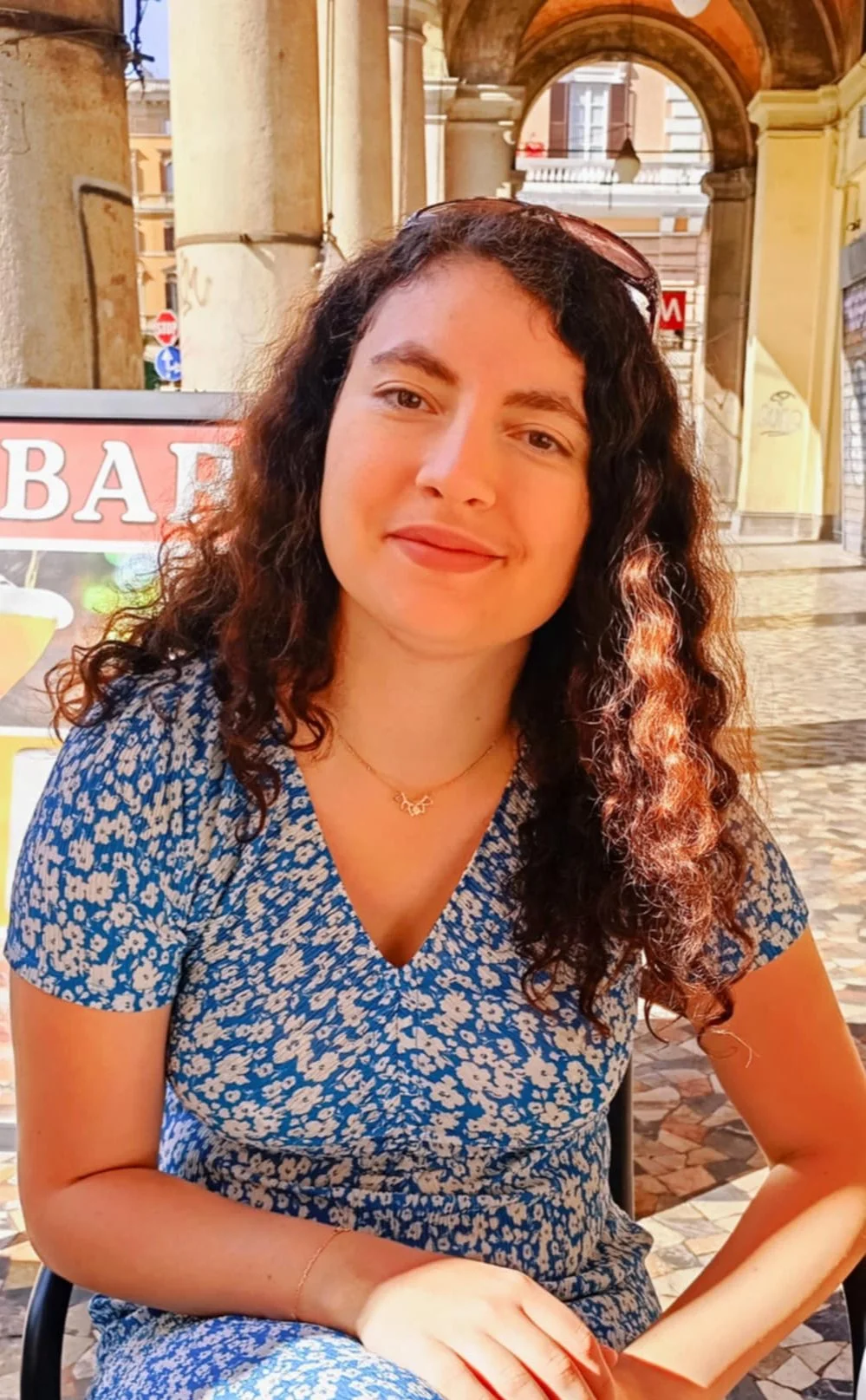 A young woman with long, curly brown hair sitting outdoors in a city street. She is wearing a blue floral dress, a butterfly necklace, and a gold bracelet. The background shows a building archway, a sign with partially visible text, and cobblestone pavement.