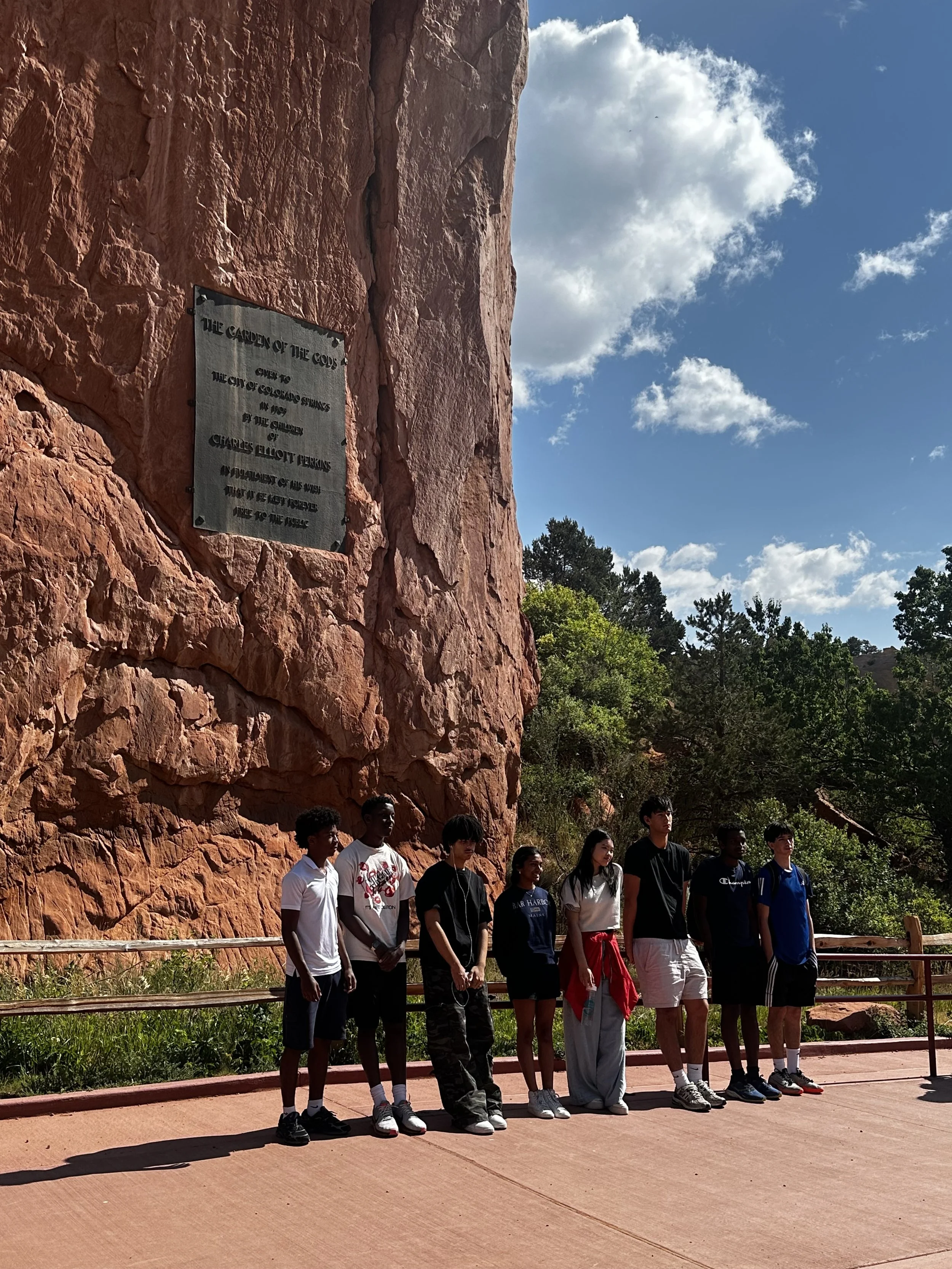 Group of teenagers standing in front of a red rock canyon wall with a plaque, trees, and clouds in the background.