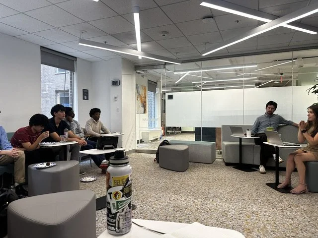 Group of people seated in a modern office or lounge area, some with laptops, facing two people who are speaking. The room has contemporary lighting and decor, with a glass partition in the background.
