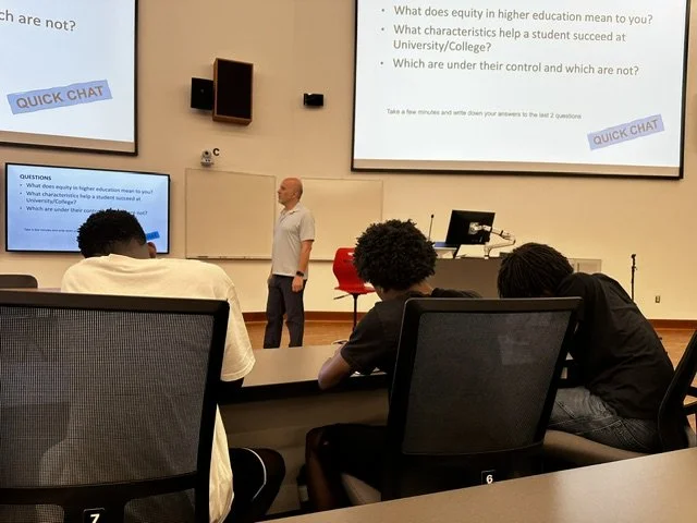 A classroom or lecture hall where a man is standing at the front near a whiteboard and two large screens; students are seated in chairs facing the instructor.