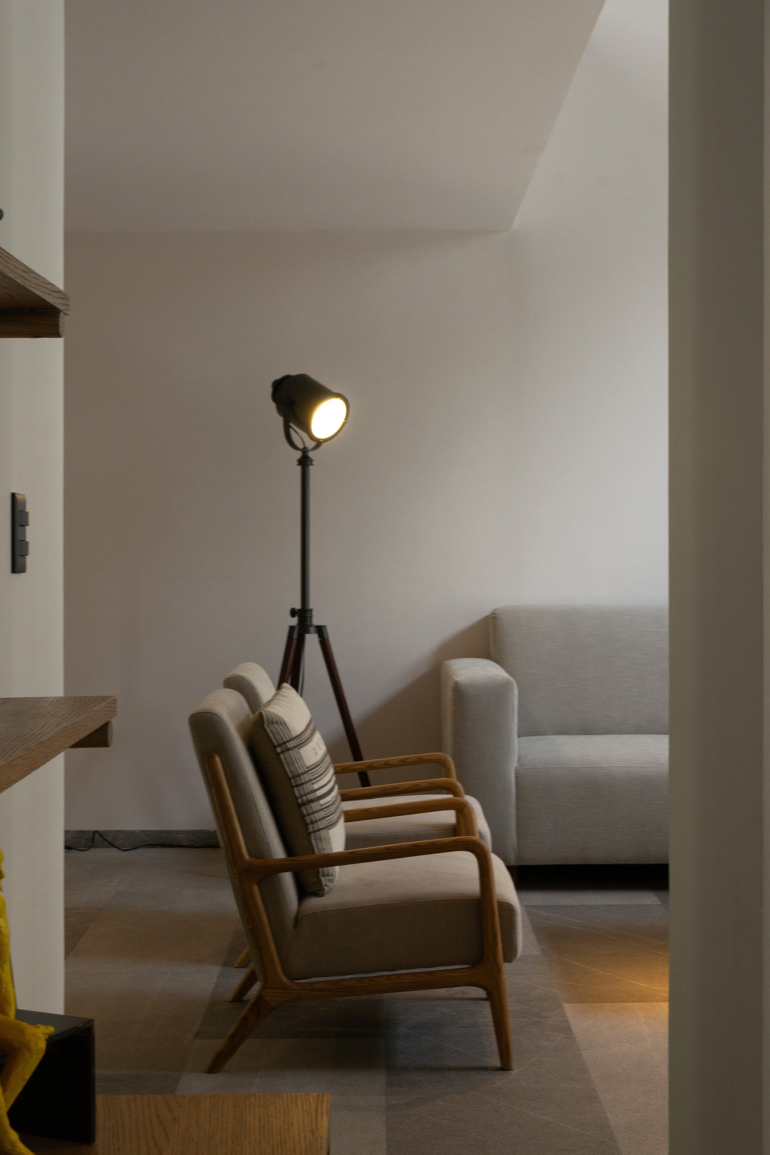 Sala de estar con sillones de madera y cojines, lámpara de pie encendida, sofá gris y pared blanca.
