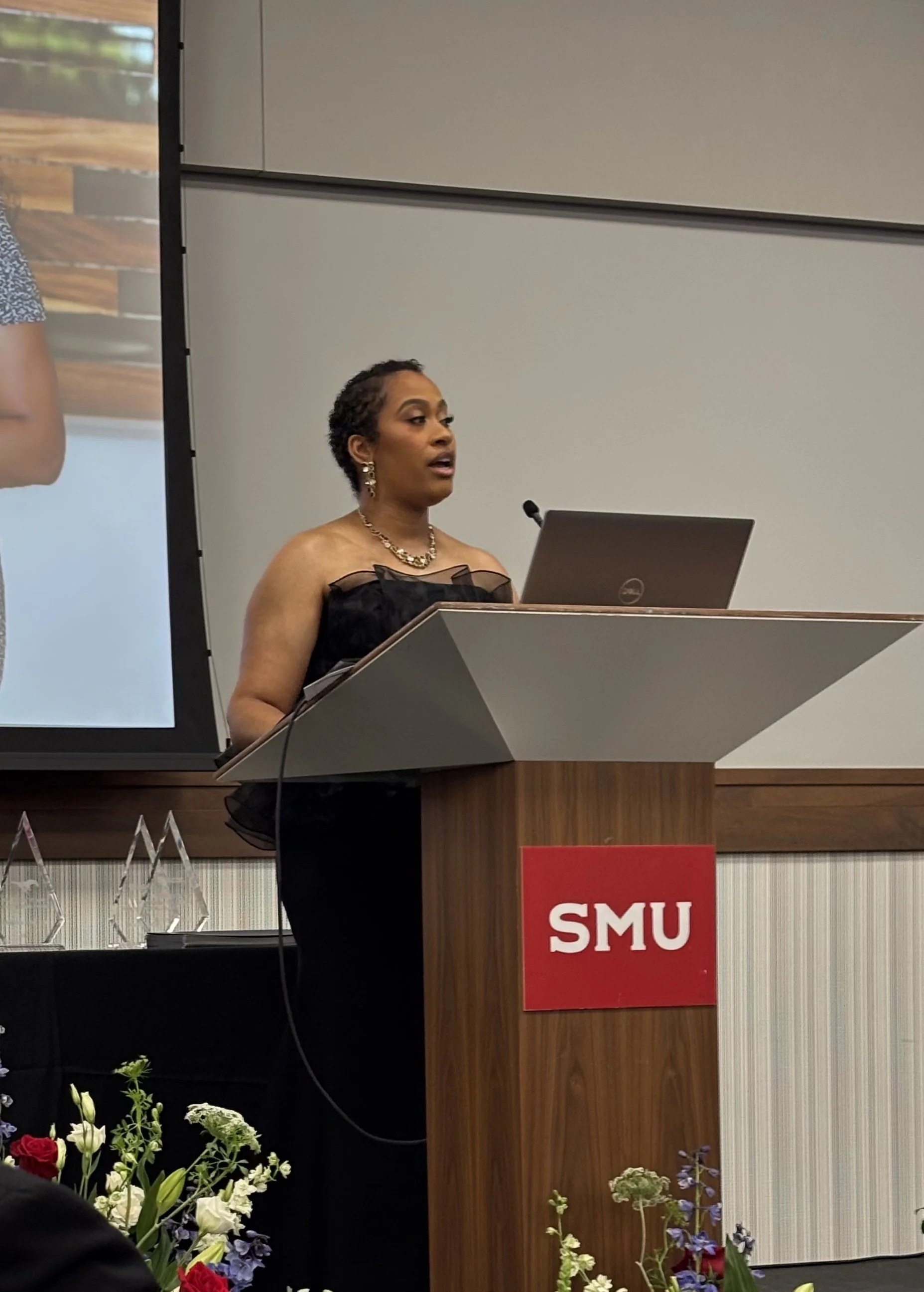 A woman speaking at a podium with on-campus flowers in the foreground and a laptop on the podium. The podium has a red sign with the letters 'SMU'.
