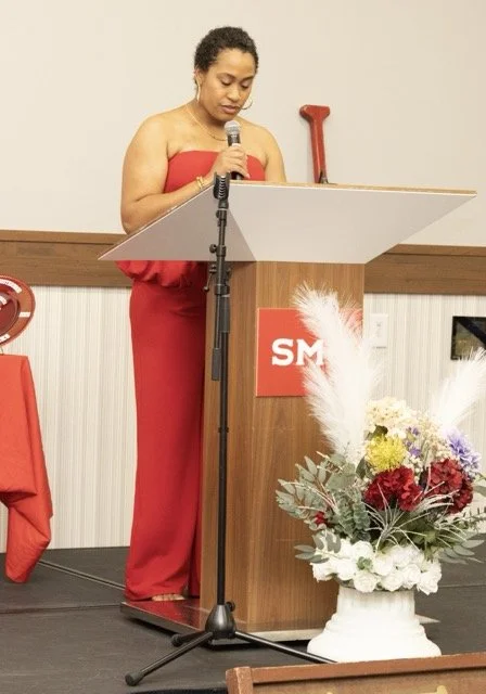 Woman in a red strapless dress standing at a podium with a microphone, reading or speaking, in a decorated room with flowers.