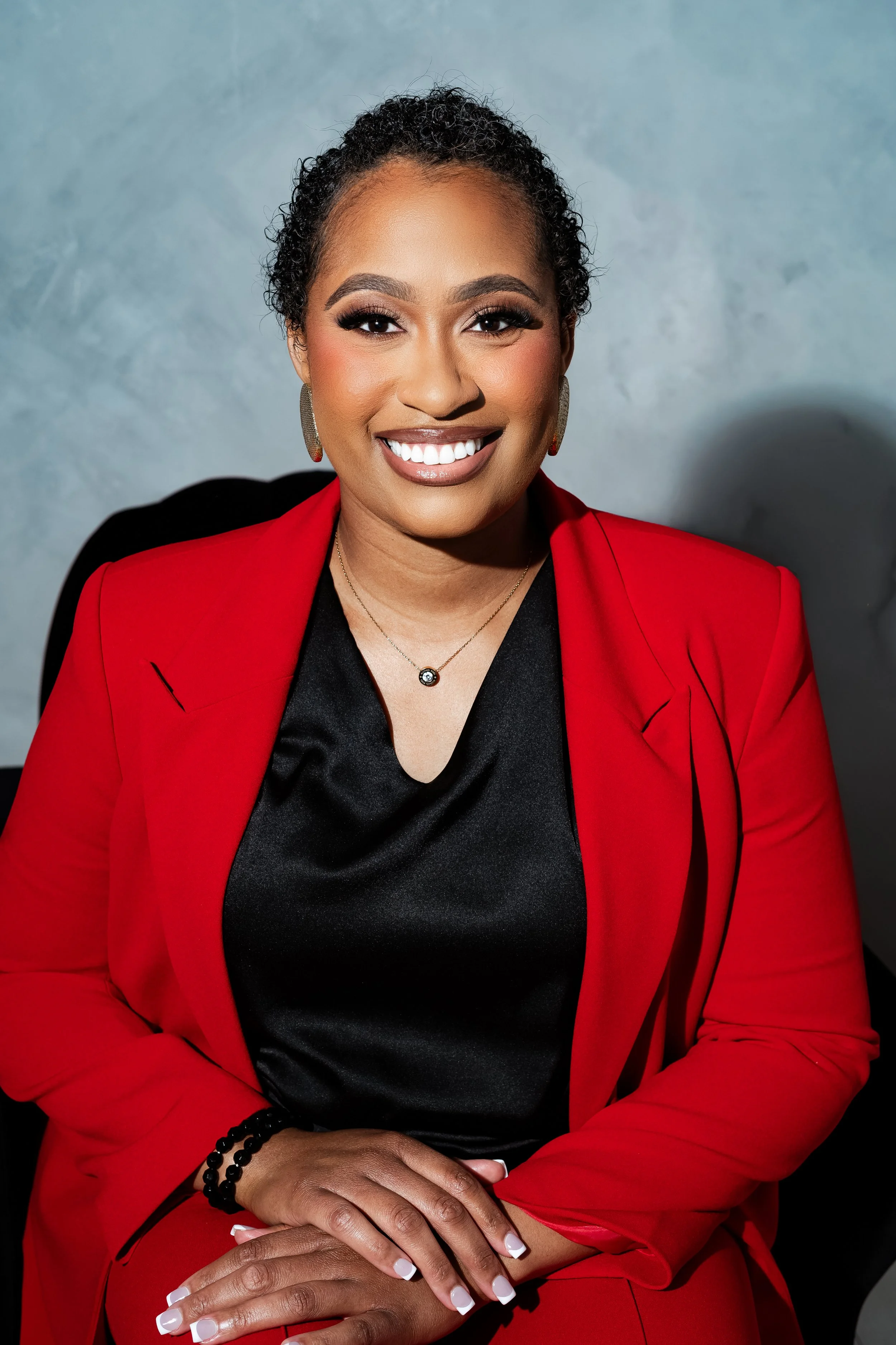 A woman wearing a red blazer and black shirt, smiling with white teeth, sitting with hands clasped. She has earrings, a necklace with a gemstone, and a black beaded bracelet, posed against a plain grey background.