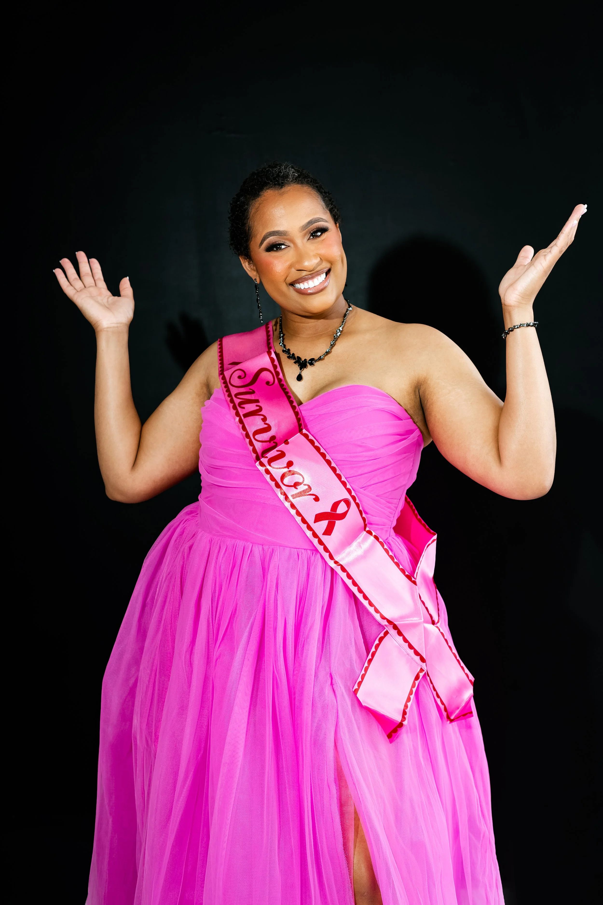 A woman in a pink strapless gown with a sash that says "Survivor" and a breast cancer awareness ribbon, smiling with her hands raised against a black background.