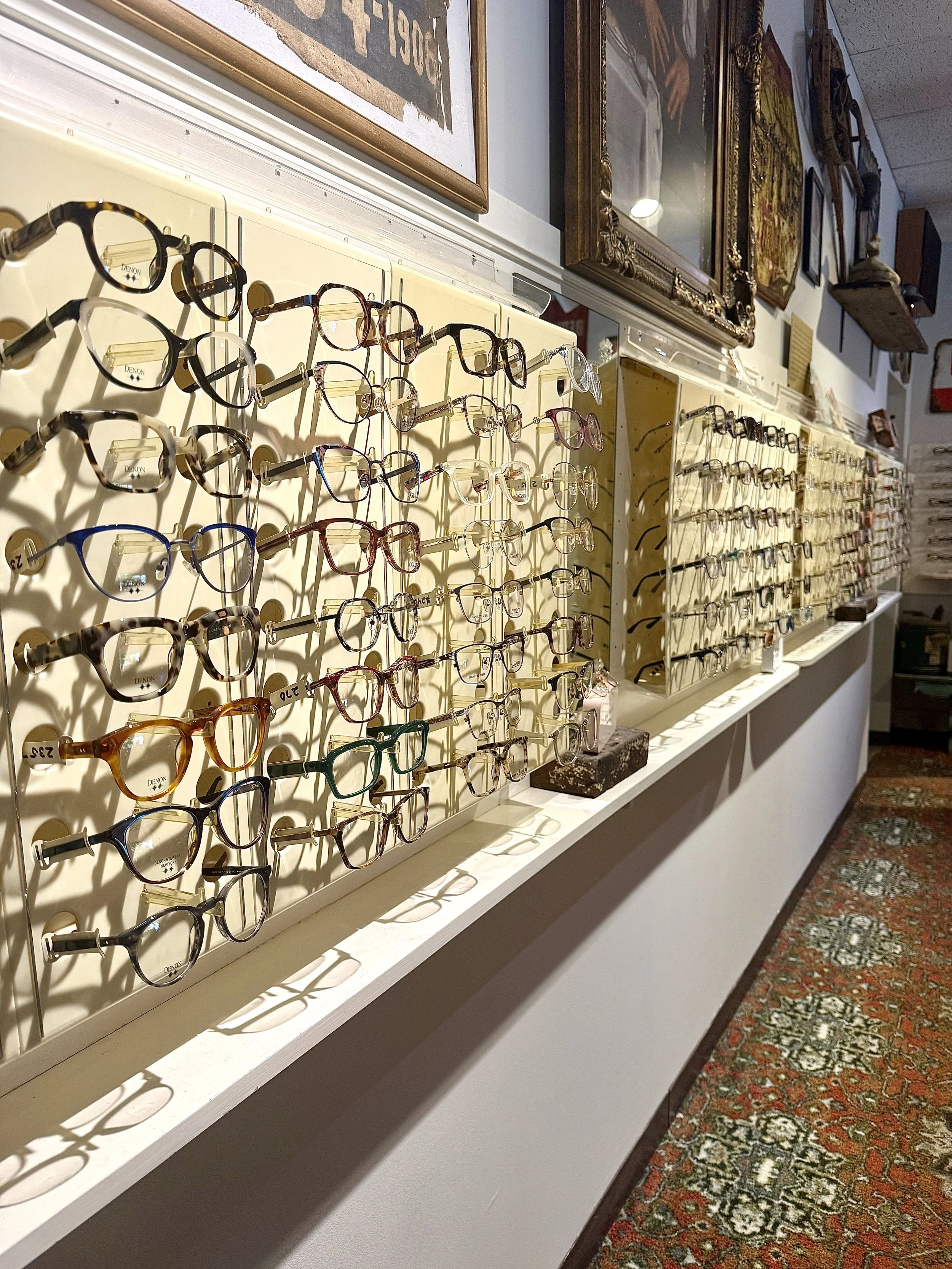 Display of various eyeglasses on a wall in an optometry store, with framed artwork and a patterned carpet visible in the background.