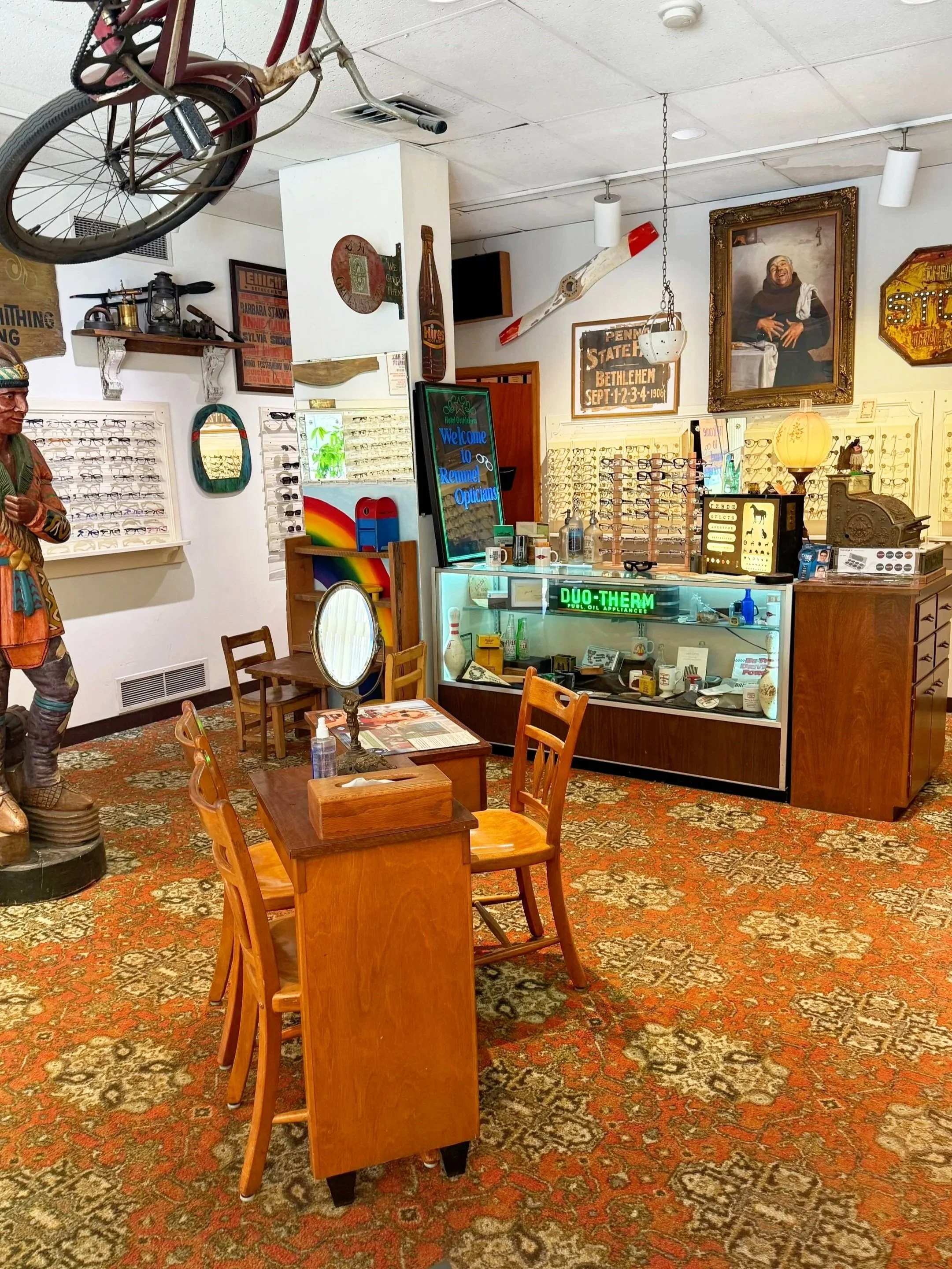 Interior of a vintage eyeglasses shop with wooden furniture, an ornate rug, displays of glasses, a neon sign that says 'Welcome to Remuda Opticians,' and various decorative items including a large portrait, a sign for Pennsylvania State Bethlehem, and a vintage bicycle and skateboard hanging from the ceiling.