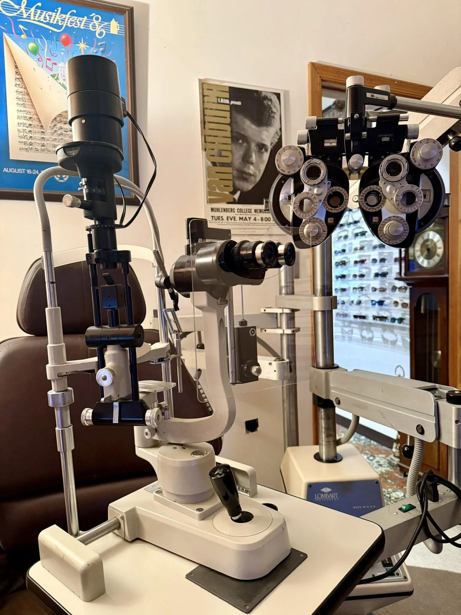 An optometry examination room contains an ophthalmic device, a slit lamp, on a white desk, with a control panel and a joystick. There are framed posters on the walls, one with a portrait of a young man and text related to a college event, and another with musical notes and text about a music festival. In the background, a display case with multiple pairs of glasses is visible.