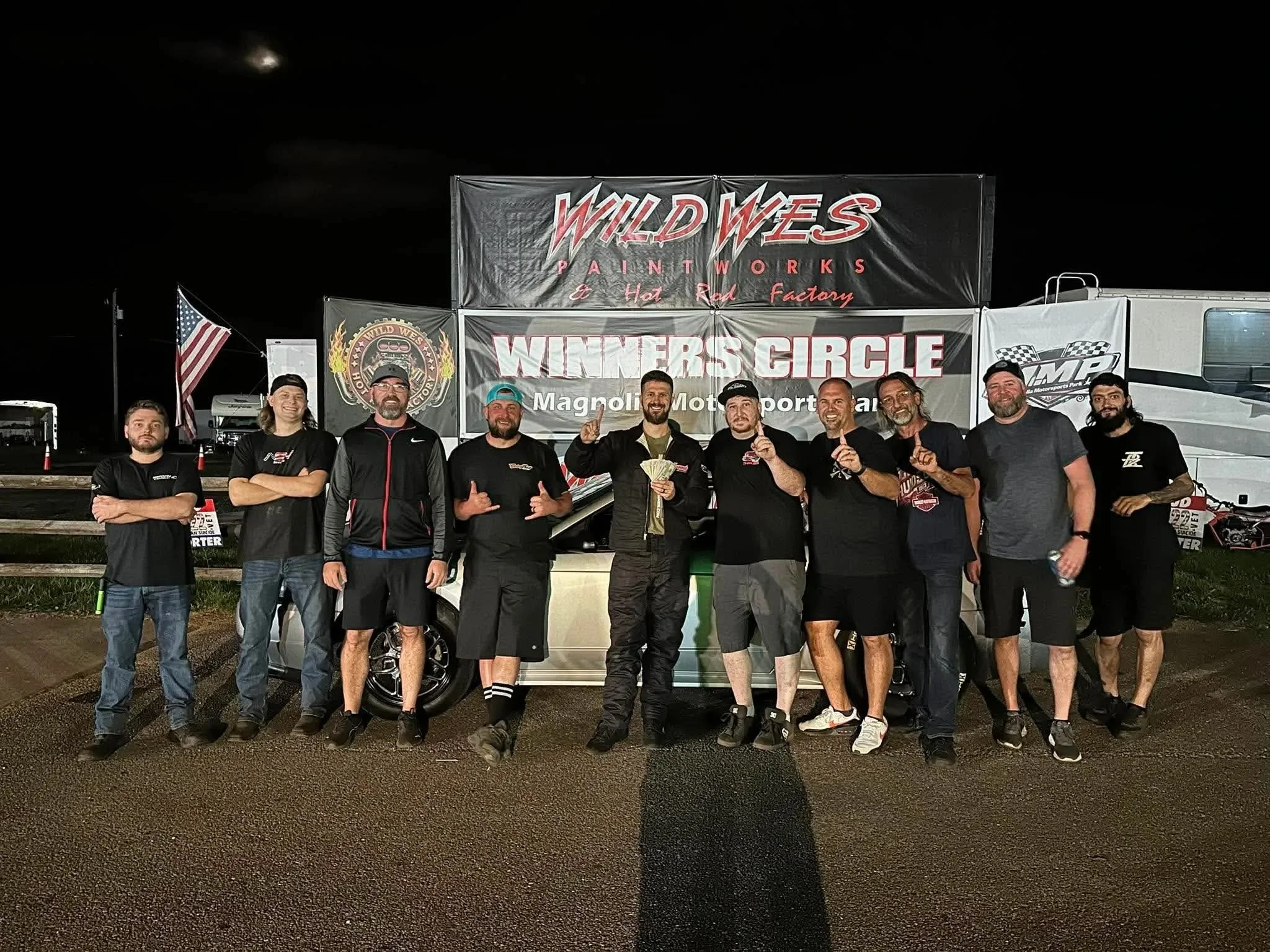 A group of ten men standing in front of a large sign that reads "Wild West Paint Works & Hot Rod Factory" and "Winners Circle." They are at night and smiling, some making gestures, with trucks and an American flag visible in the background.