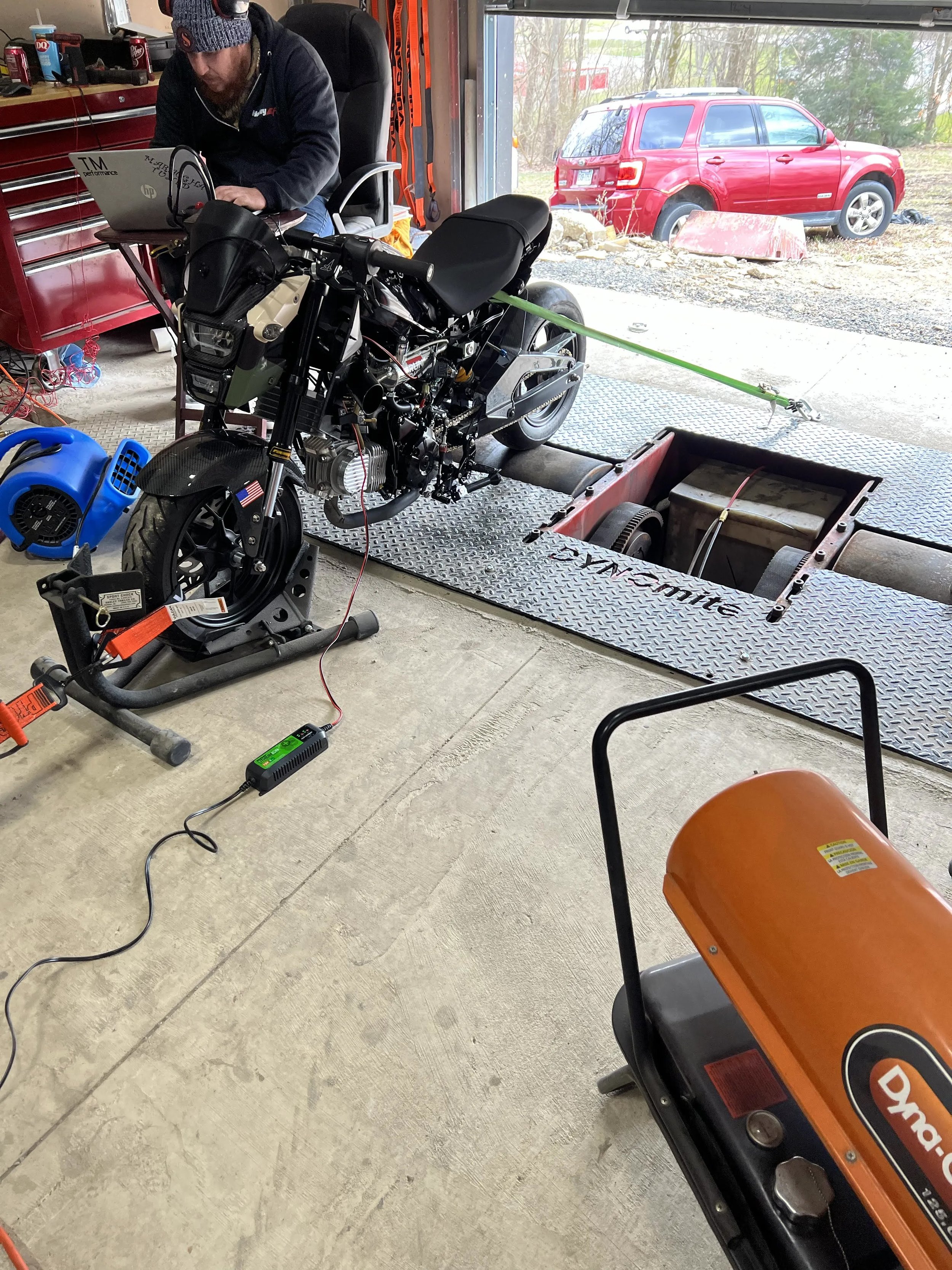 A man working on a motorcycle in a garage, with tools and equipment around him. Outside the garage, there is a red SUV parked on the driveway.
