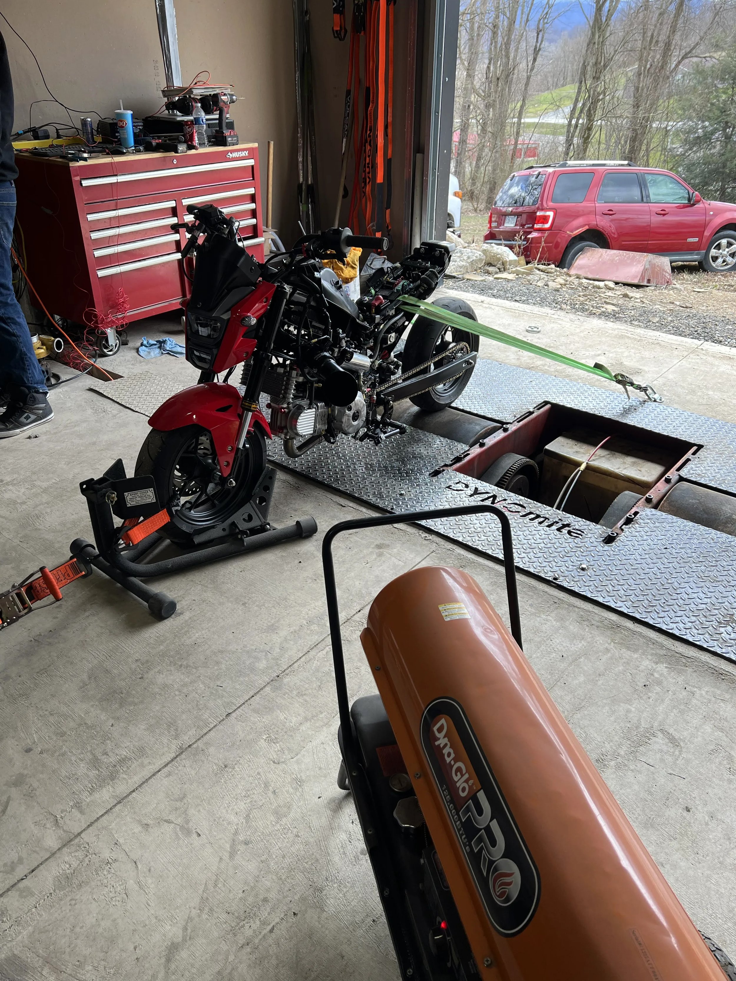 A garage interior with a motorcycle on a dyno testing stand, various tools in a red Husky tool chest, and a view of outside through the open garage door with a red SUV parked nearby.